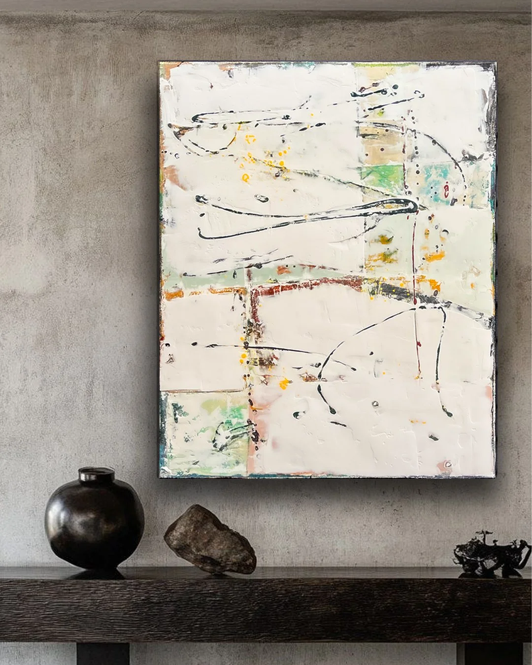 Abstract painting with squiggly black lines, splashes of yellow, orange, green, and red, mounted on a concrete wall above a dark wooden shelf with a black round vase, a rock, and a small sculpture of a motorcycle.