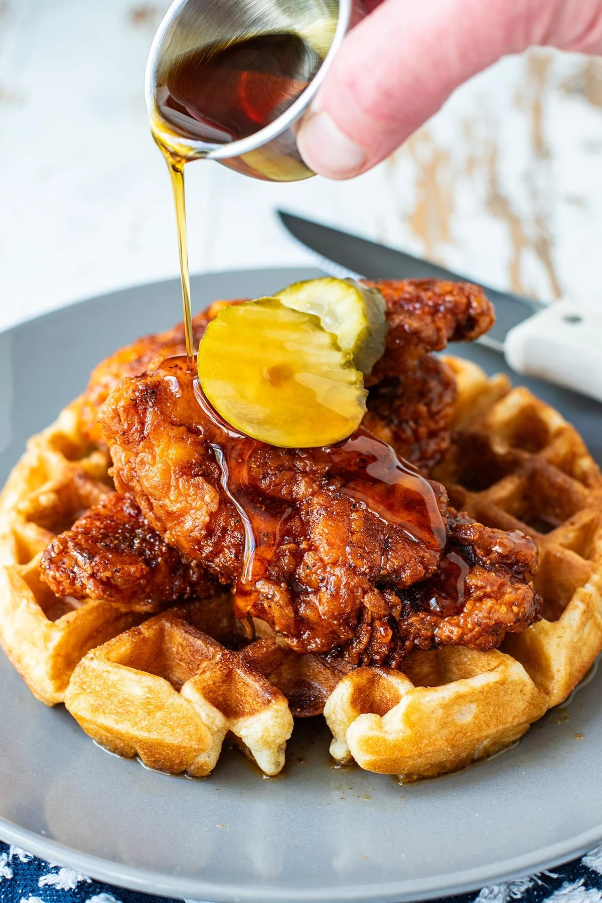 Close-up of a waffle topped with fried chicken, with syrup and a pickle slice being poured on top.