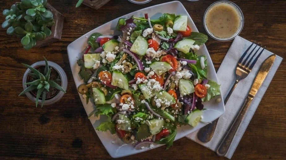 Fresh salad with mixed greens, cherry tomatoes, cucumbers, red onions, olives, crumbled feta cheese, and a side of salad dressing on a wooden table.