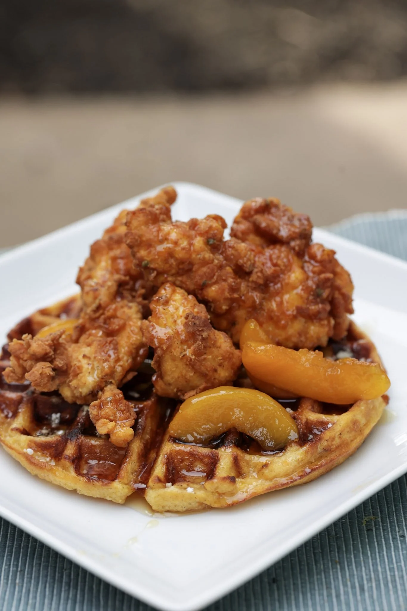Close-up of a waffle topped with fried chicken, caramelized peaches, and syrup, served on a white plate.