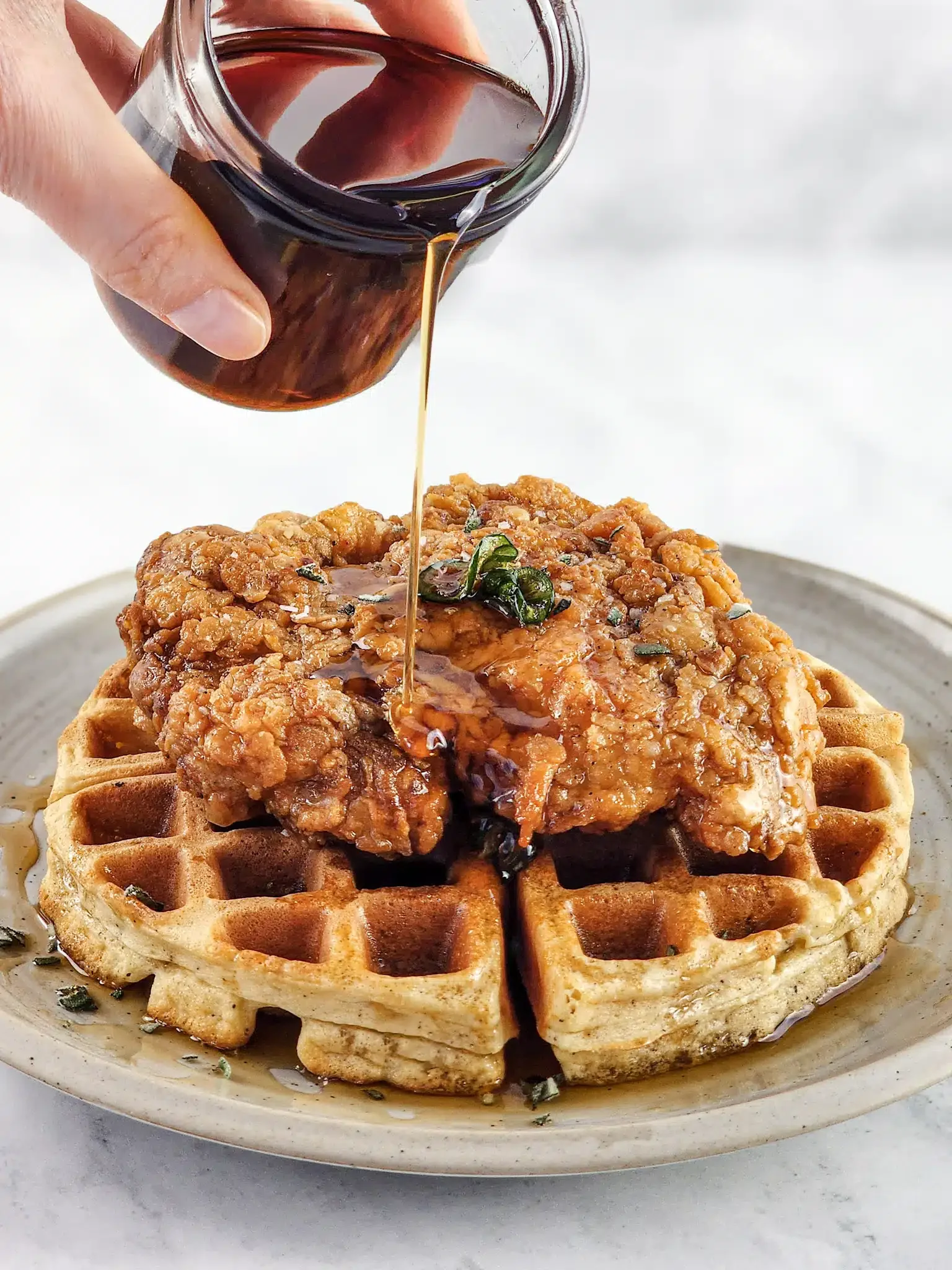 A plate with a waffle topped with a fried chicken drumstick, and syrup being poured from a glass container.
