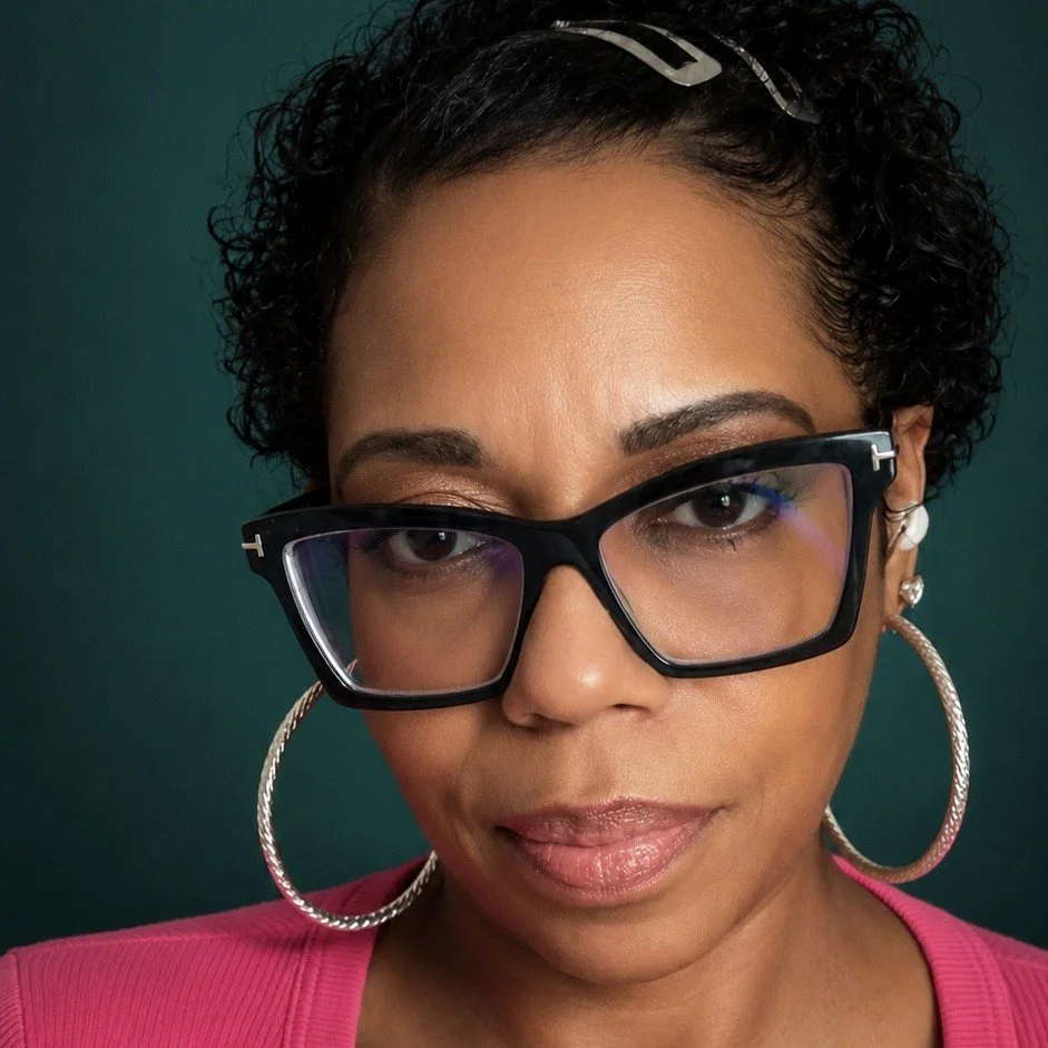 Close-up of a woman with short curly hair wearing large hoop earrings and black glasses, dressed in a pink top, against a dark background.