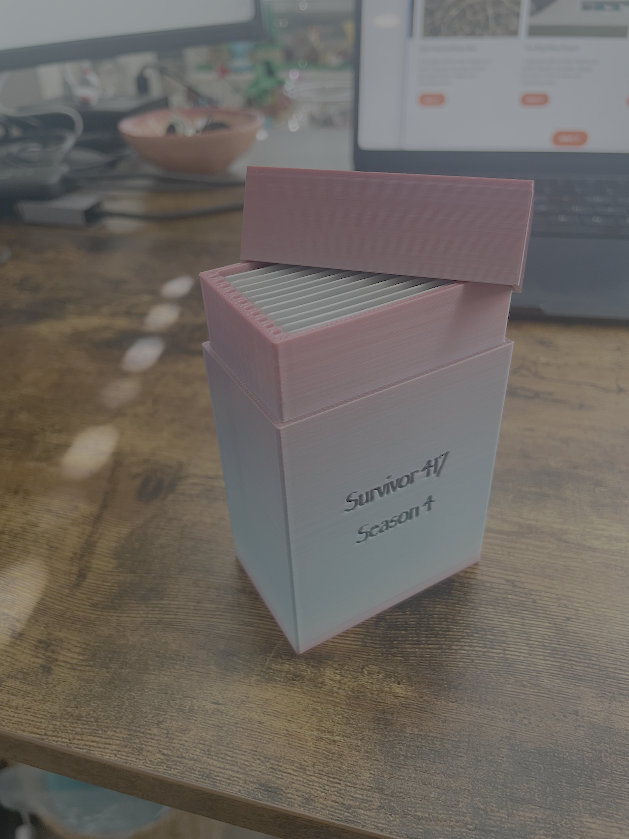 A pink and white box labeled "Survivor47 Season 4" on a wooden table, with a laptop and a bowl in the blurred background.