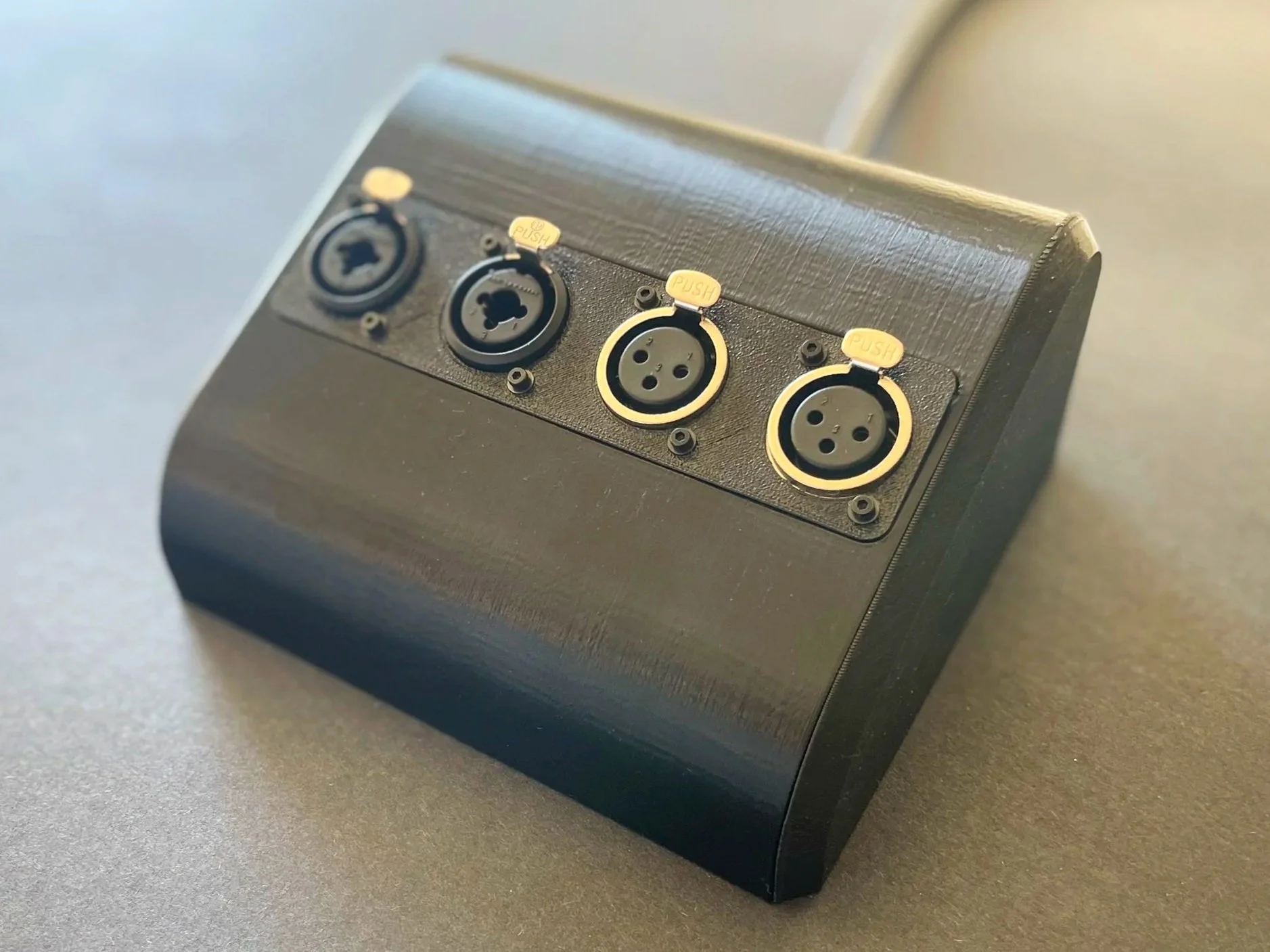 A power strip with four outlet ports on a dark surface, near a clear glass with a blue straw and a plastic container, with a blurred background of a kitchen or workspace.