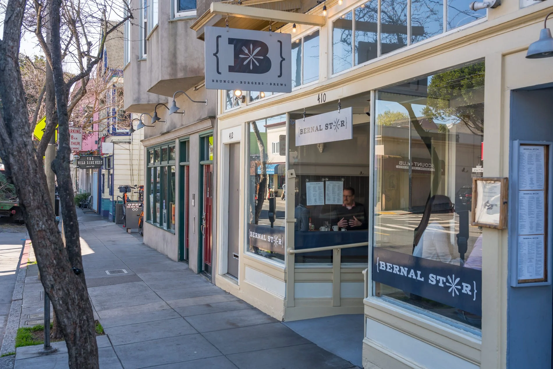 Street view of a small business storefront with signs that read 'Brunch Burgers Brews' and 'Bernal St'. The storefront has large glass windows and a man sitting inside near the window. The sidewalk and trees line the street, with neighboring building