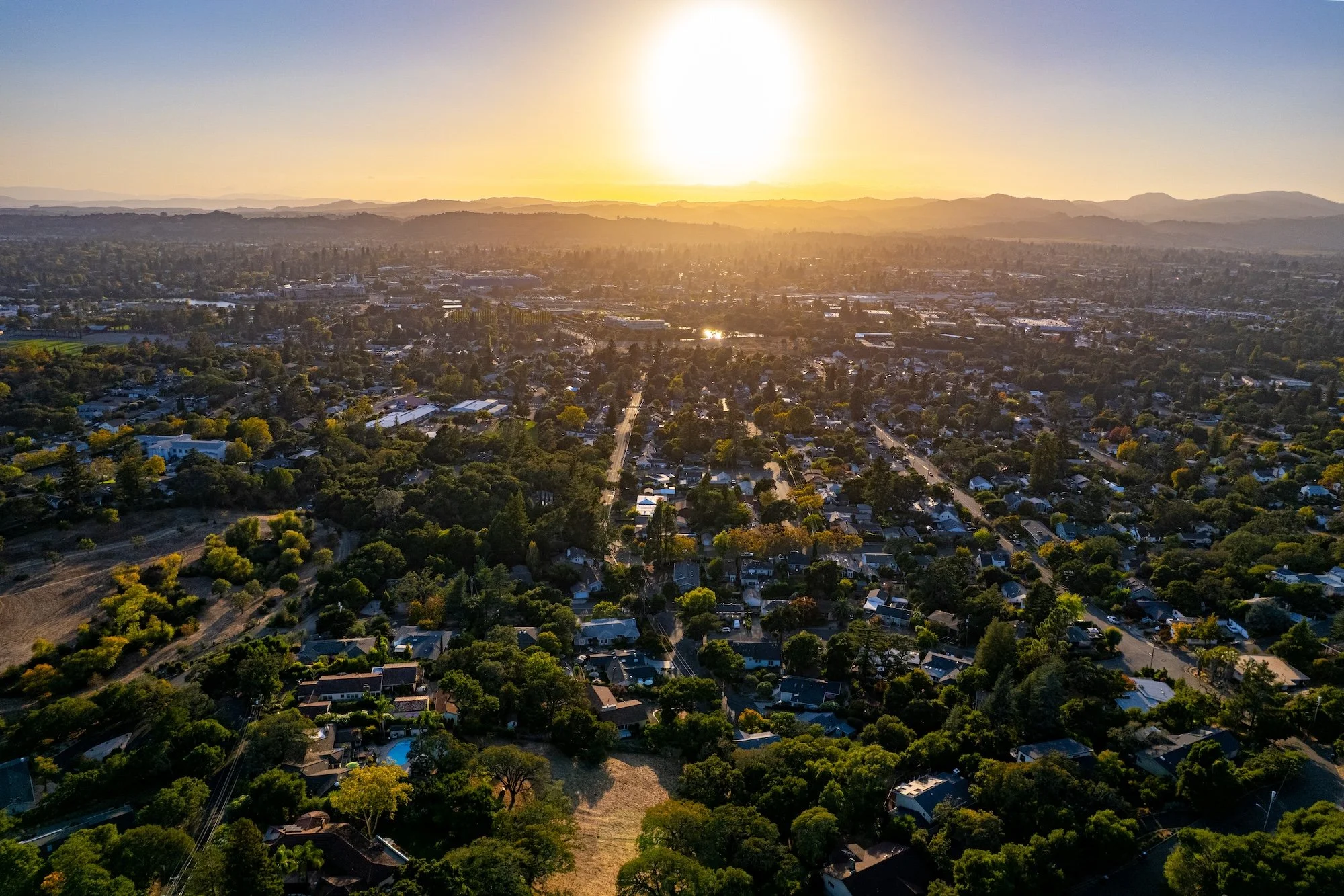 An aerial view of a residential area at sunset with houses, trees, and streets with mountains in the background.