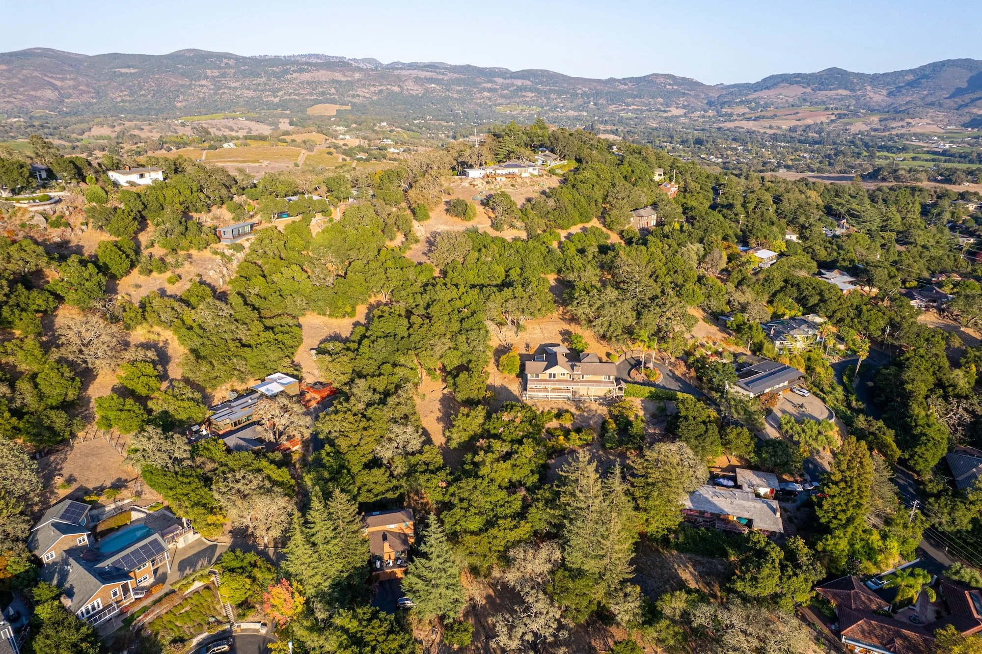 An aerial view of a hilly residential area with houses surrounded by lush greenery and trees, with a mountain range and clear blue sky in the background.