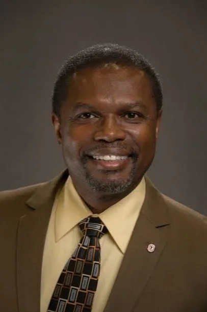 Portrait of a smiling man with short black hair and a beard, wearing a brown suit, beige shirt, and patterned tie, against a dark gray background.
