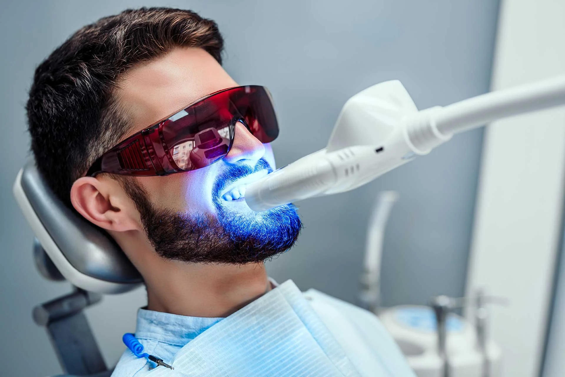 A man with dark hair and a beard undergoing a dental procedure using a blue light with protective glasses on.