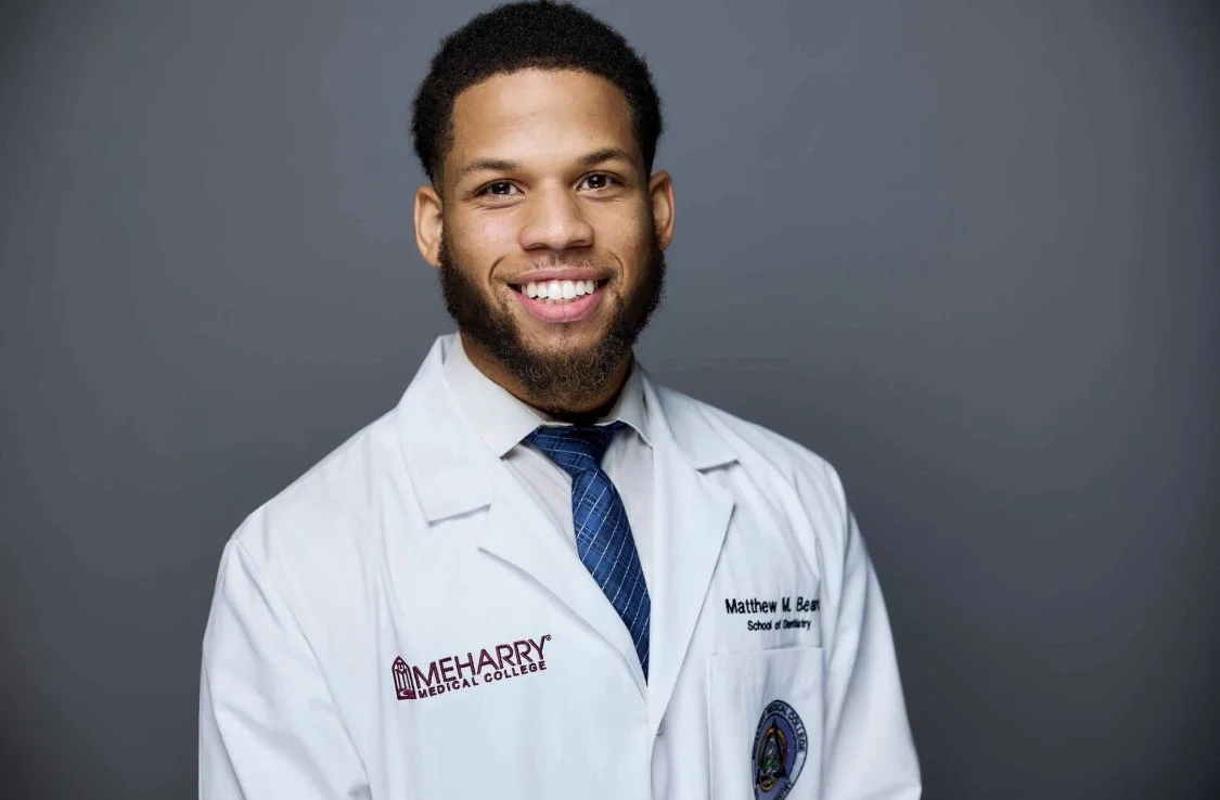A young African American man in a white medical coat with his name and institution embroidered, smiling against a gray background.