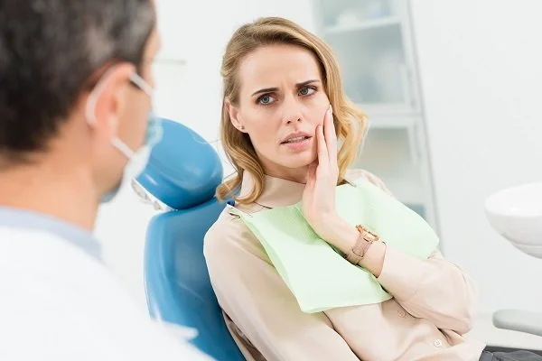 A woman sitting in a dental chair, holding her jaw with a worried expression, while a dentist examines her.