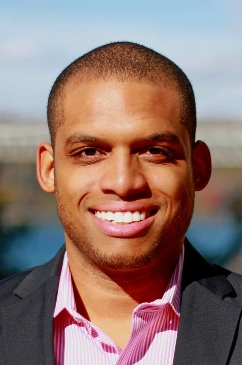 A smiling young man with short hair, wearing a dark blazer and a pink striped shirt, standing outdoors with a blurred background.