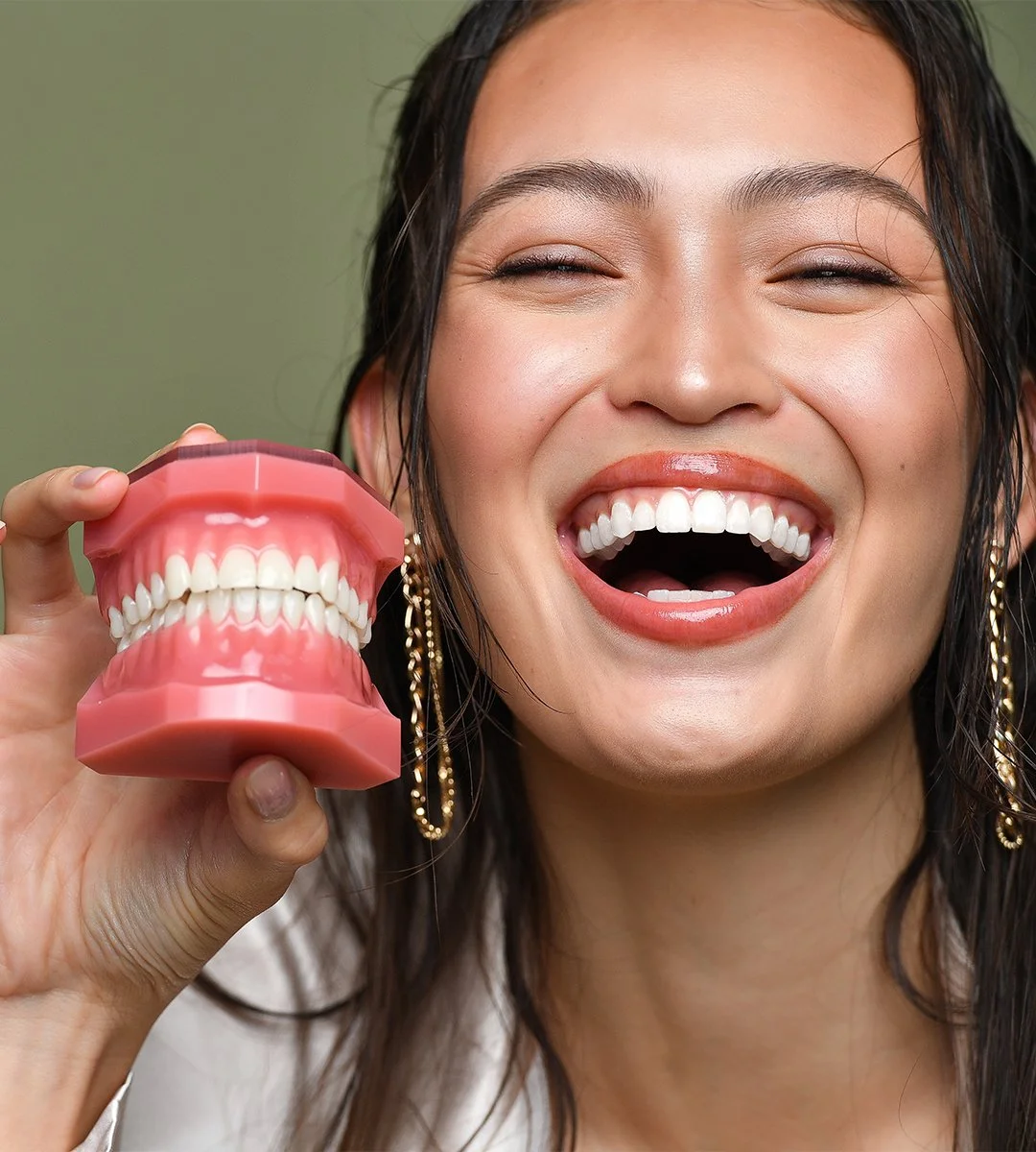 A woman with wet hair smiling and holding a large model of human teeth and gums.