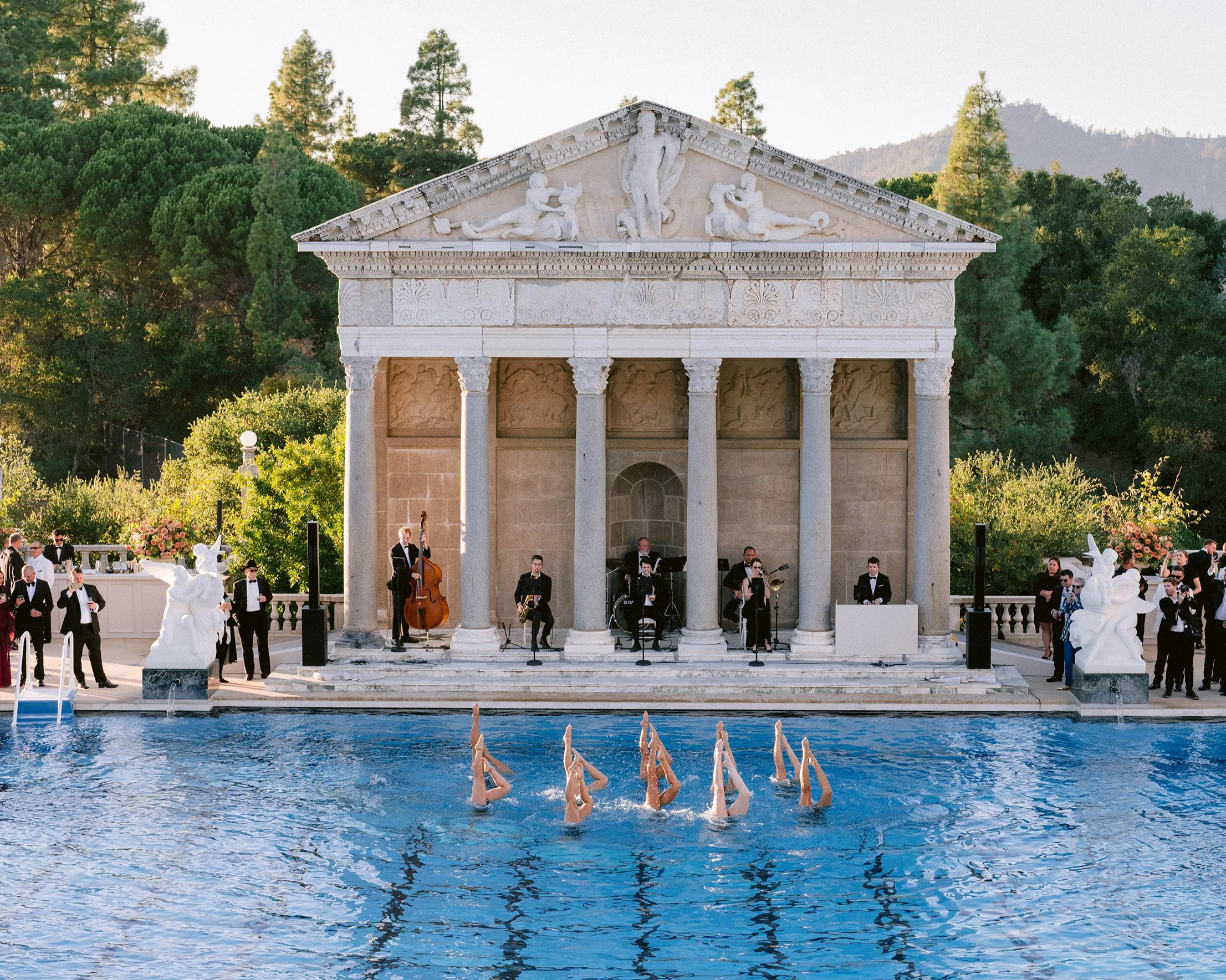 A classical Greek-style temple with a detailed frieze and sculptures on the pediment, set in a lush green landscape with mountains in the background. In front, a live band performs, and a swimming pool with synchronized dancers' legs extended above t