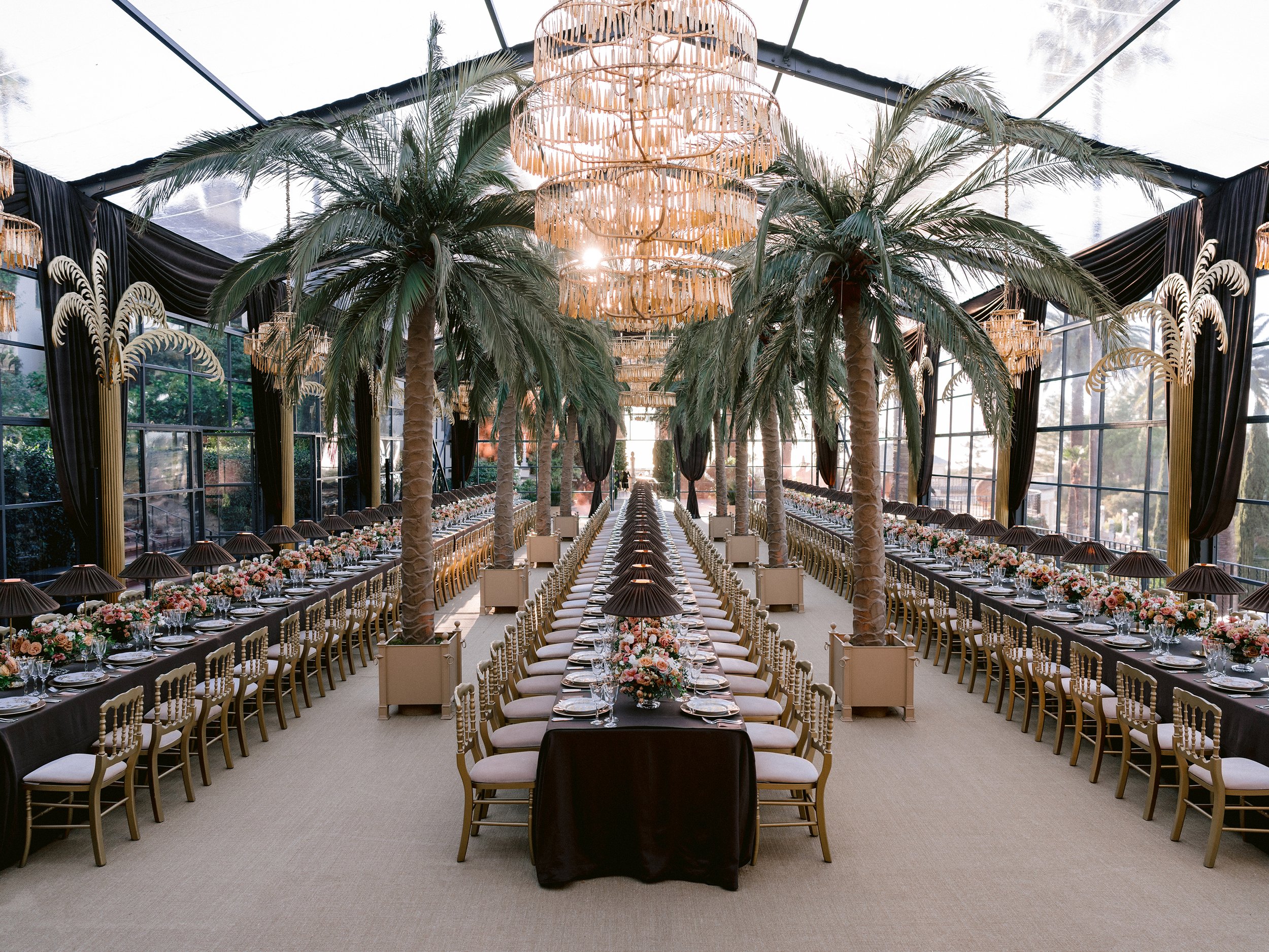 Elegant banquet hall decorated with tall palm trees, black drapery, and large chandeliers for a formal event or wedding reception.