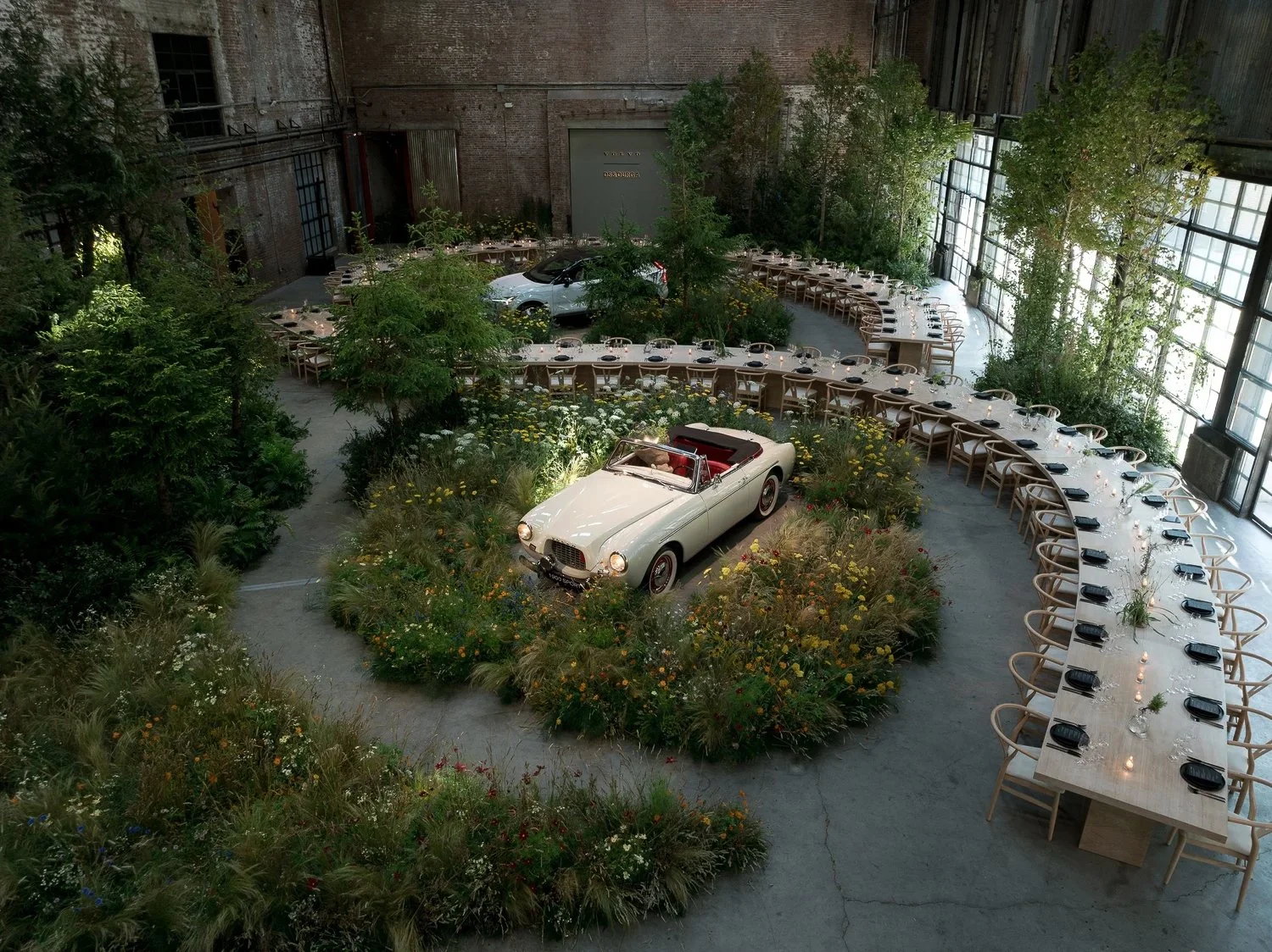 An indoor event space featuring a vintage white convertible car and a classic silver SUV, surrounded by lush greenery and arranged for a formal gathering with long tables set with black napkins and candles.