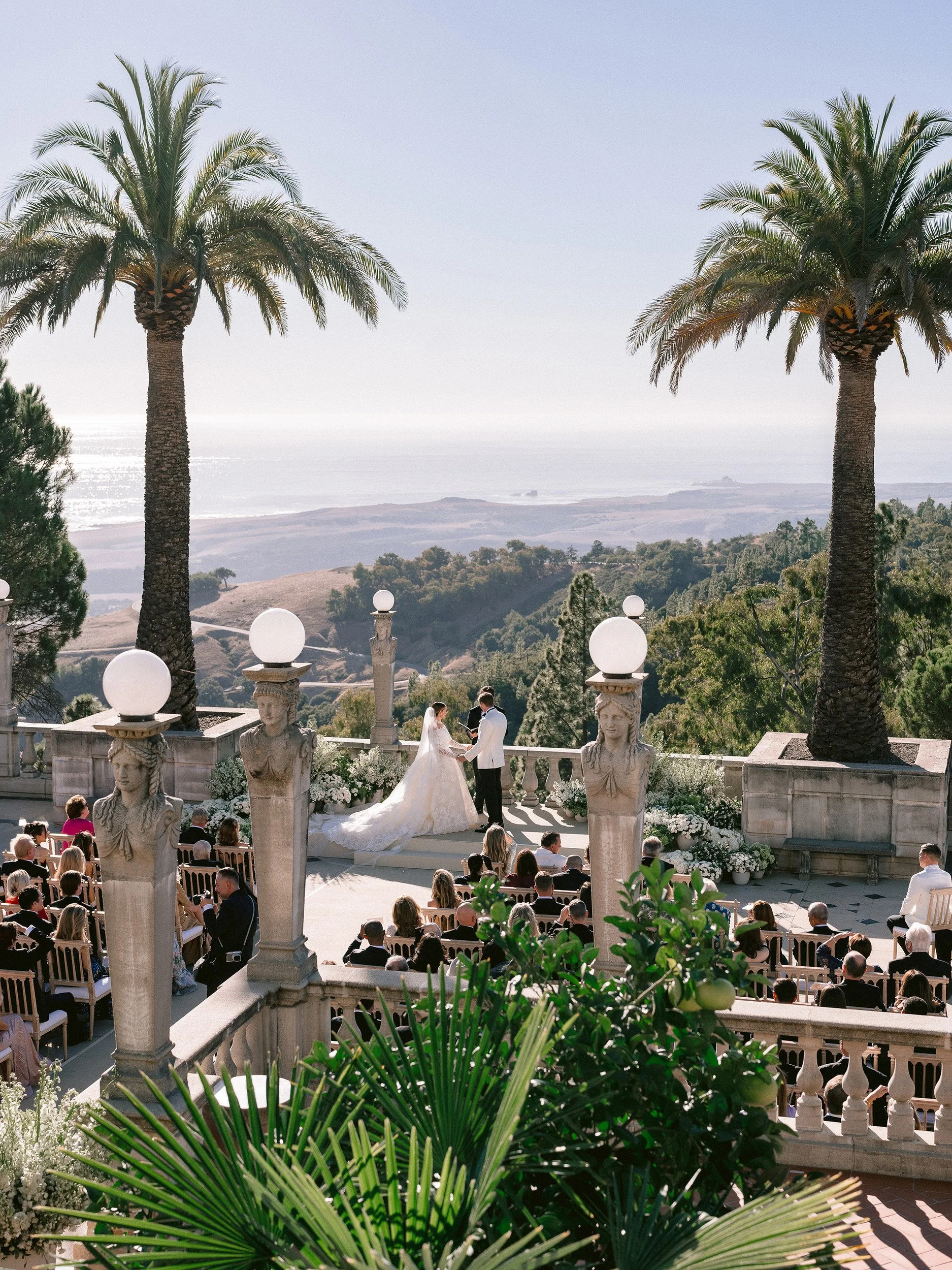 A wedding ceremony taking place outdoors on a terrace with scenic ocean and hills in the background. A bride and groom are standing in front of seated guests, exchanging vows. The setting features classical statues and large palm trees, with lush gre