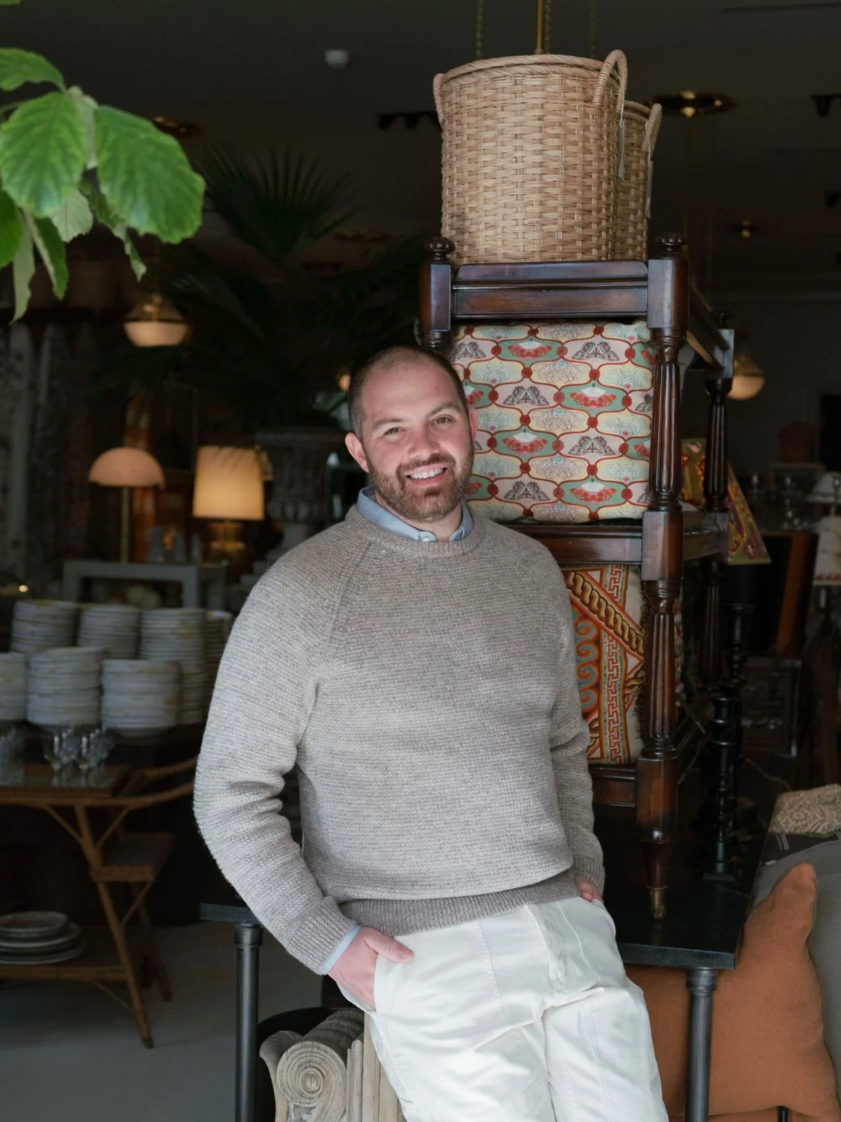 A man standing indoors in a store or showroom, smiling at the camera, with stacked chairs and decorative items behind him.