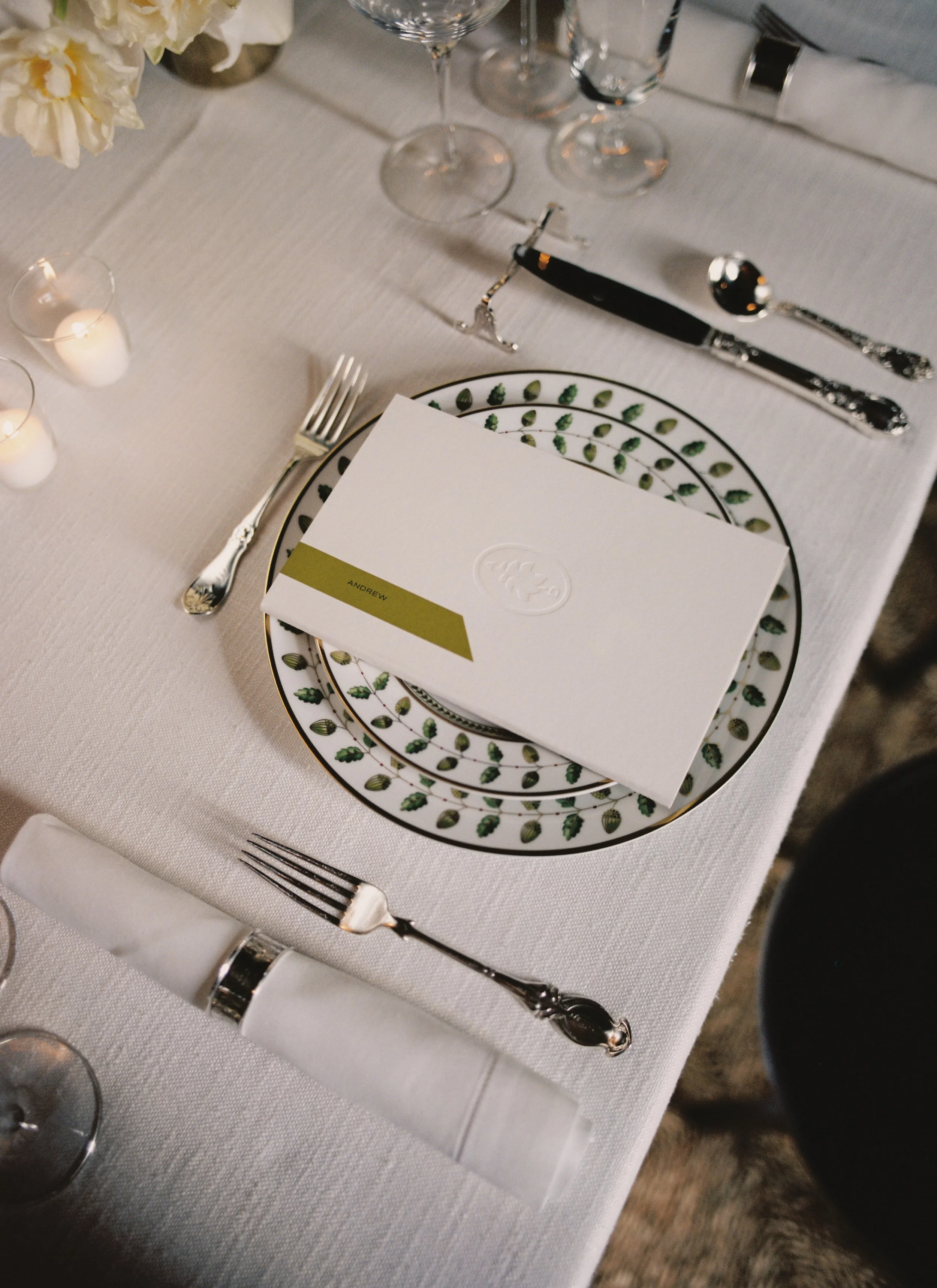 Elegant table setting with a white tablecloth, silverware, water glasses, candles, a floral centerpiece, and a place card with the name 'Andrew' on a decorative plate.