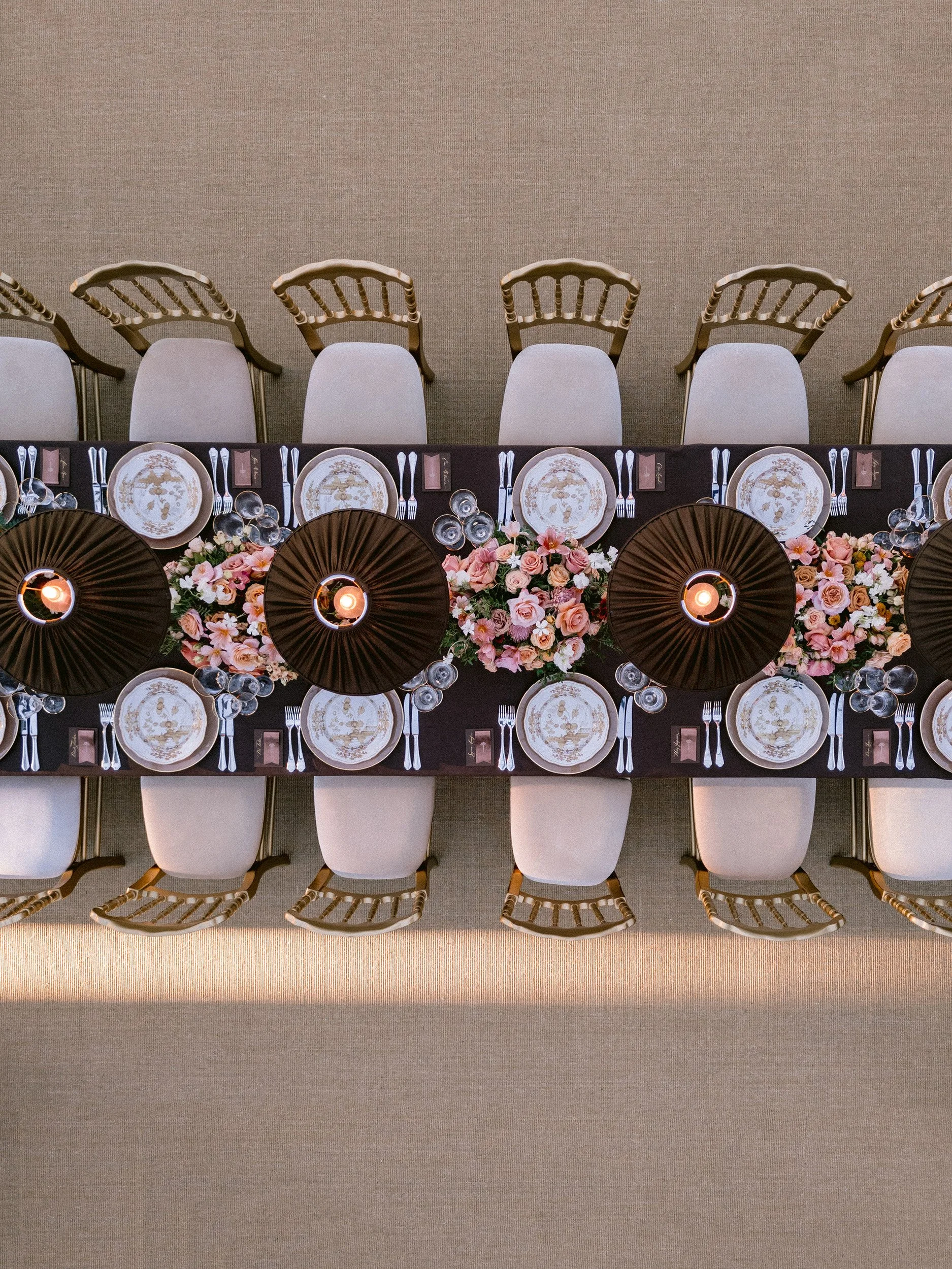 Overhead view of a long banquet table decorated with pink and white flowers, lit candles, and decorated plates, set for a formal event with white chairs surrounding it.