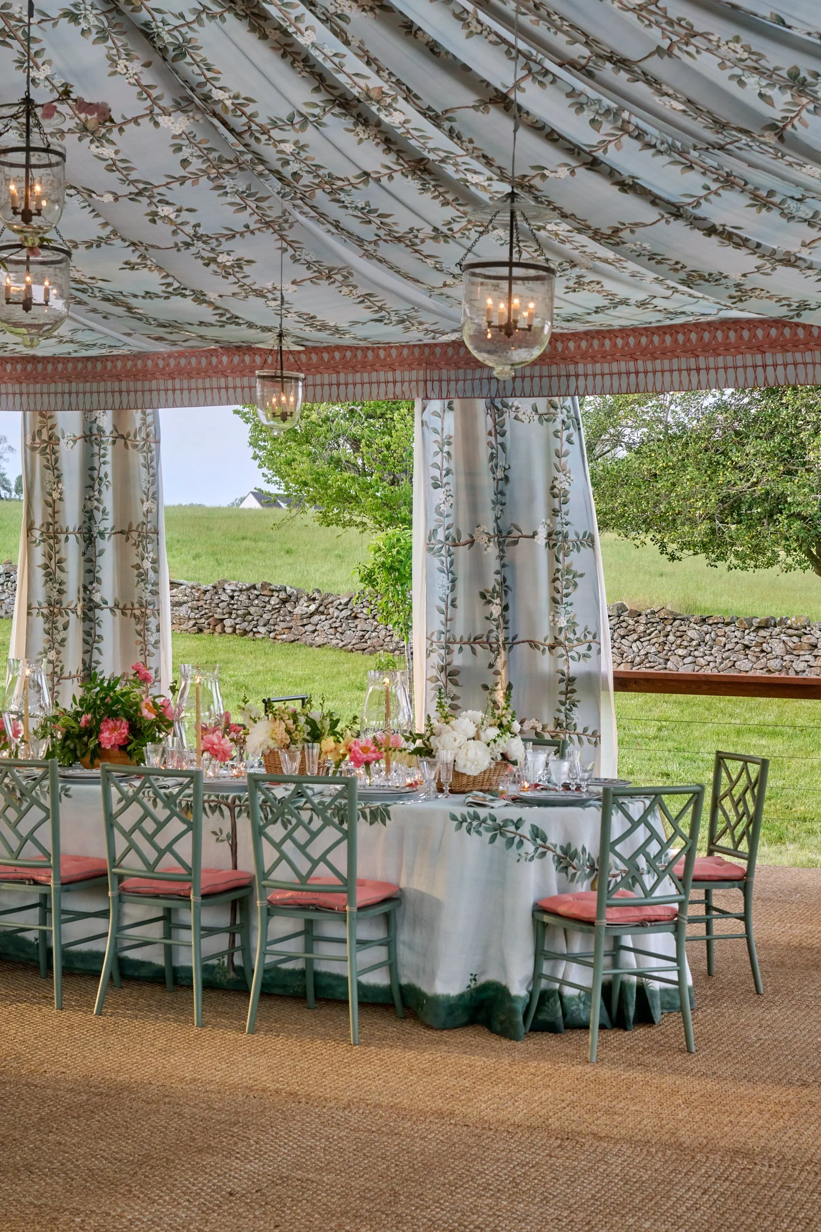 Elegant outdoor dining setup under a decorated canopy with floral and leaf patterns, featuring a long table with flowers, tableware, and chairs, with lush greenery and stone wall in the background.