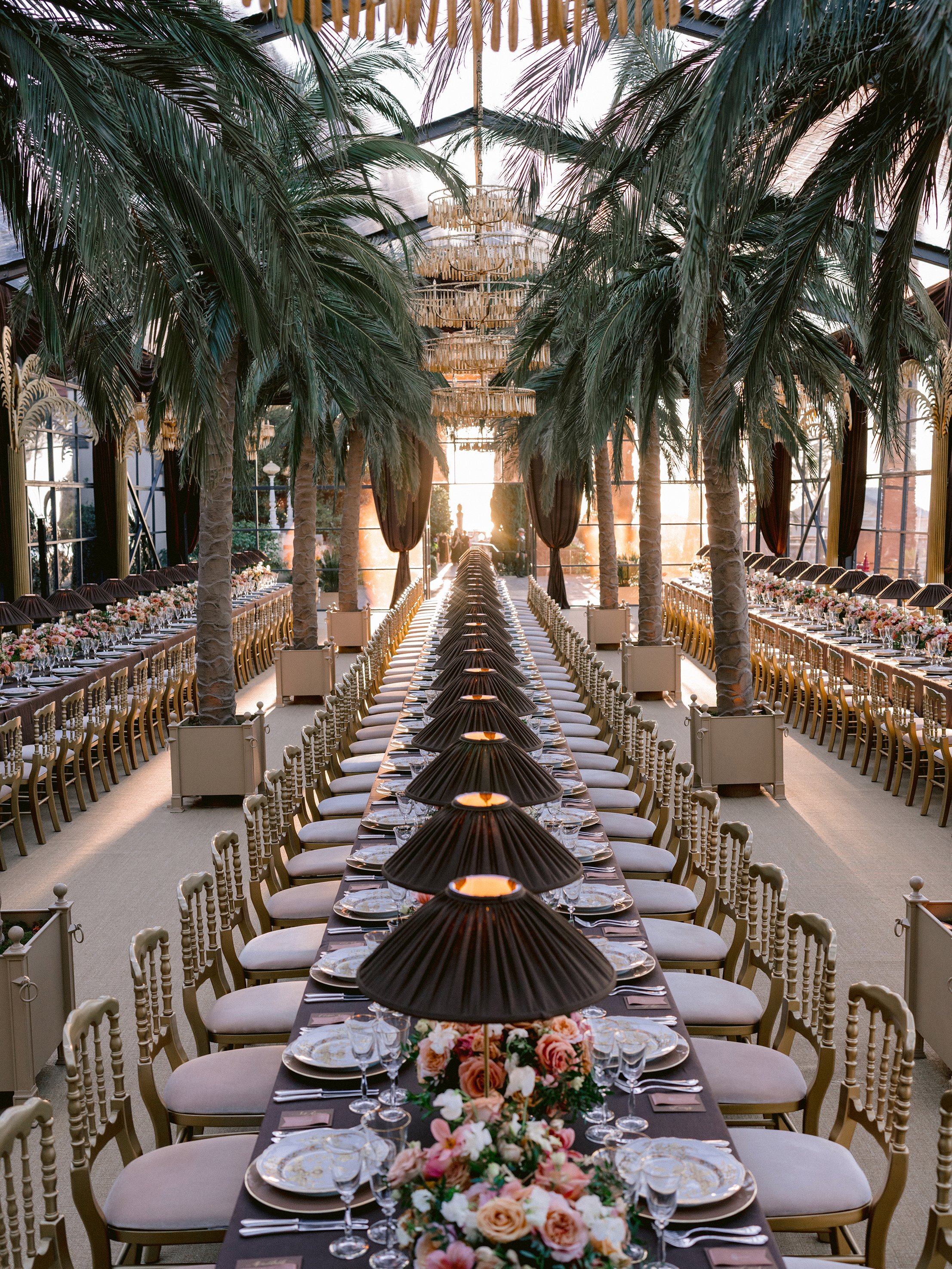 A long banquet-style table set for a formal event with elegant place settings, floral centerpieces, and overhead black fans in an indoor space with tall glass windows, lush green palm trees, and chandeliers.