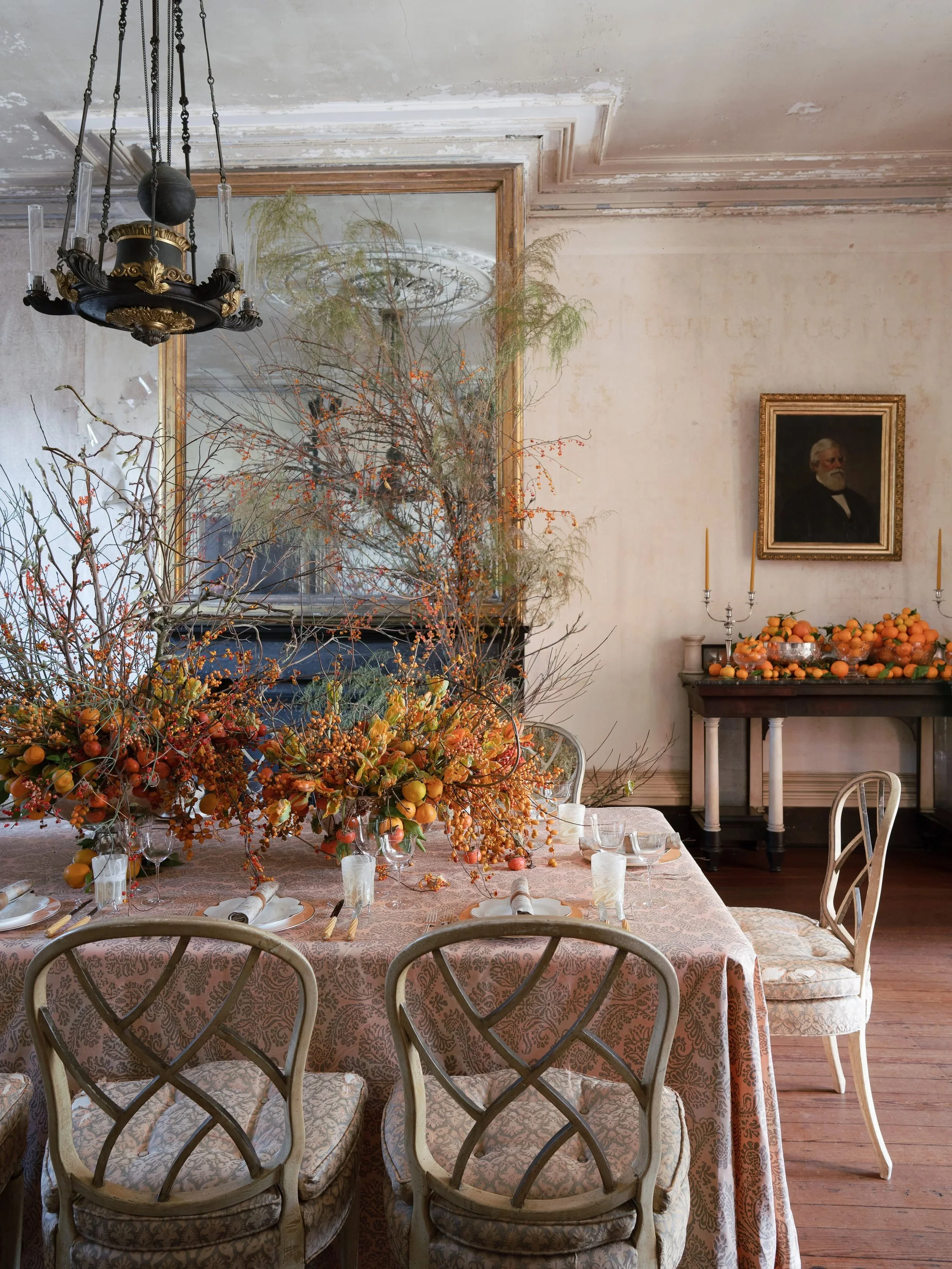 A dining room table set for a formal dinner with floral and fruit centerpieces, surrounded by elegant chairs. The room features a large mirror, a chandelier, framed portrait, and a side table with candles and orange-like fruits.