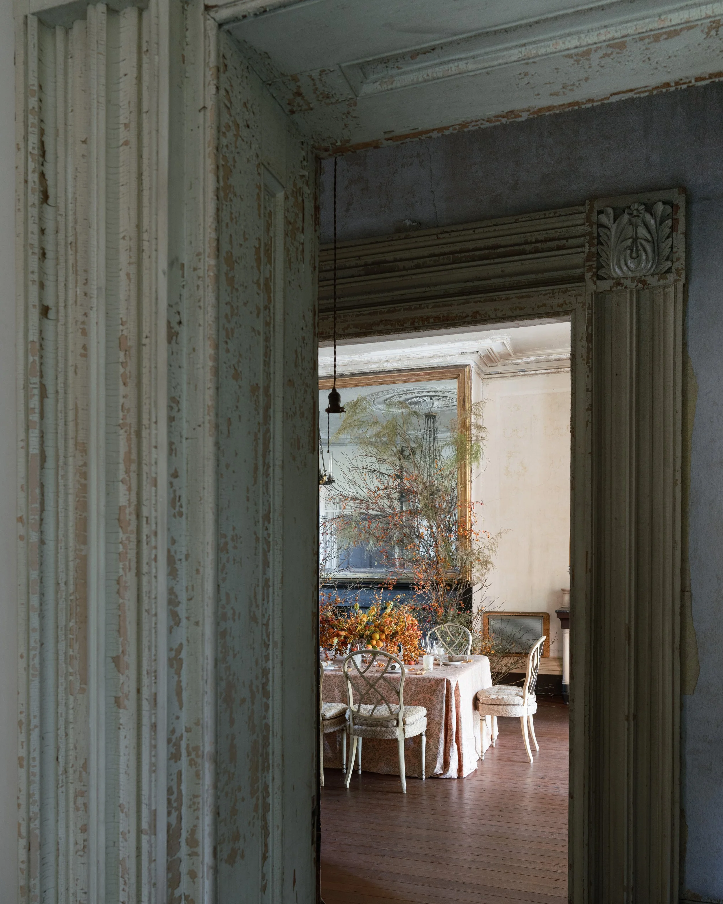 View of an elegant dining room with a large table covered with a tablecloth, decorated with a floral and fruit centerpiece. The room features high ceilings, ornate crown molding, and natural light coming through large windows. The walls are painted in a soft, warm tone.