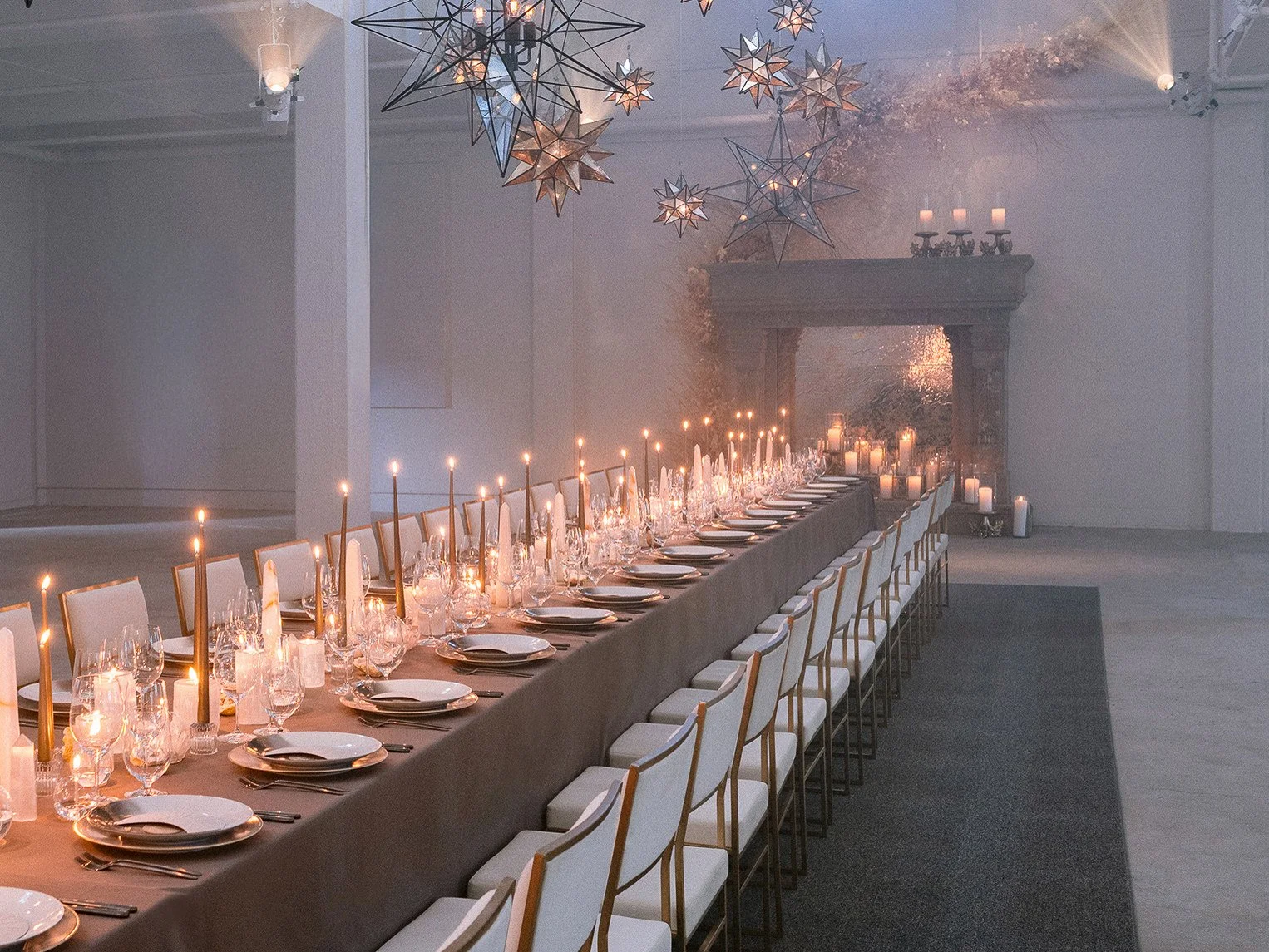 A long banquet table decorated with candles, plates, glasses, and silverware, set for a formal dinner. The room has modern star-shaped pendant lights hanging from the ceiling and a fireplace mantel with candles in the background. Candles are also pla
