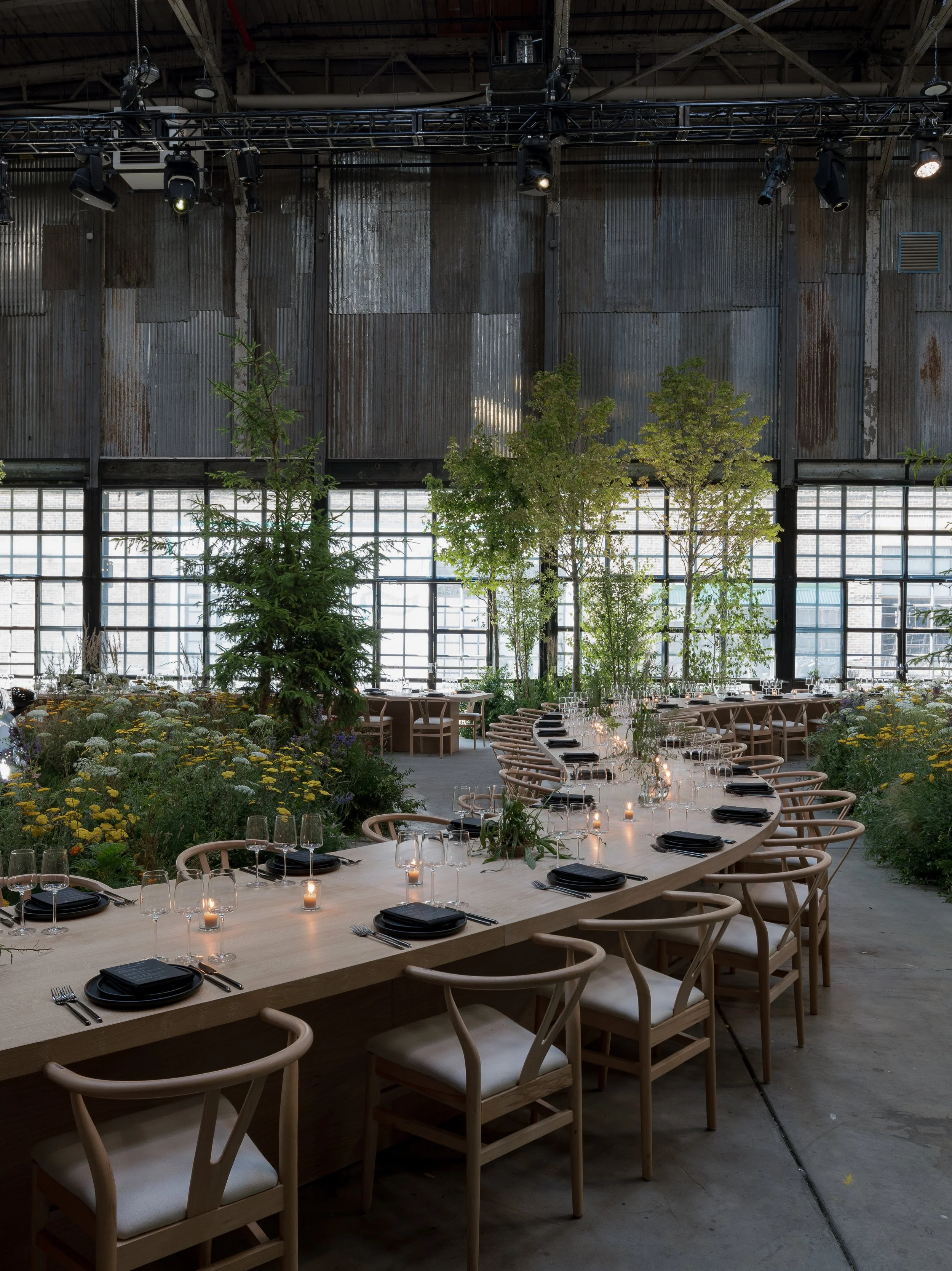 Long dining table set with black plates, glasses, and candles in an industrial-style venue with corrugated metal walls, large windows, and greenery.
