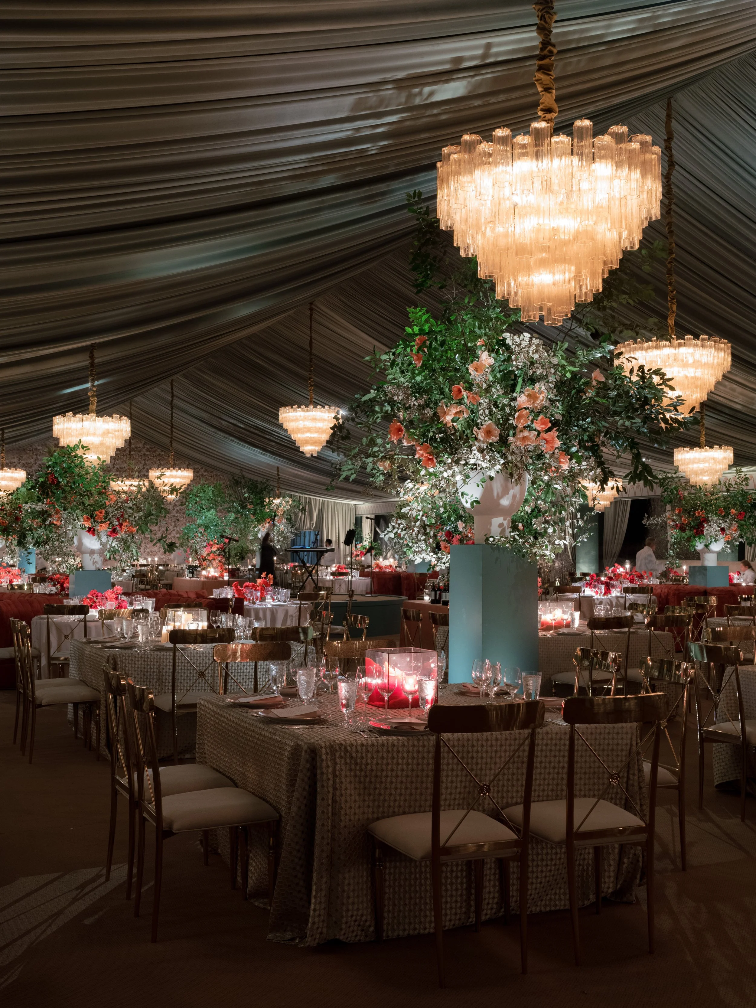 Elegant banquet hall with large floral centerpieces, crystal chandeliers, round tables with formal place settings, and draped ceiling fabric.