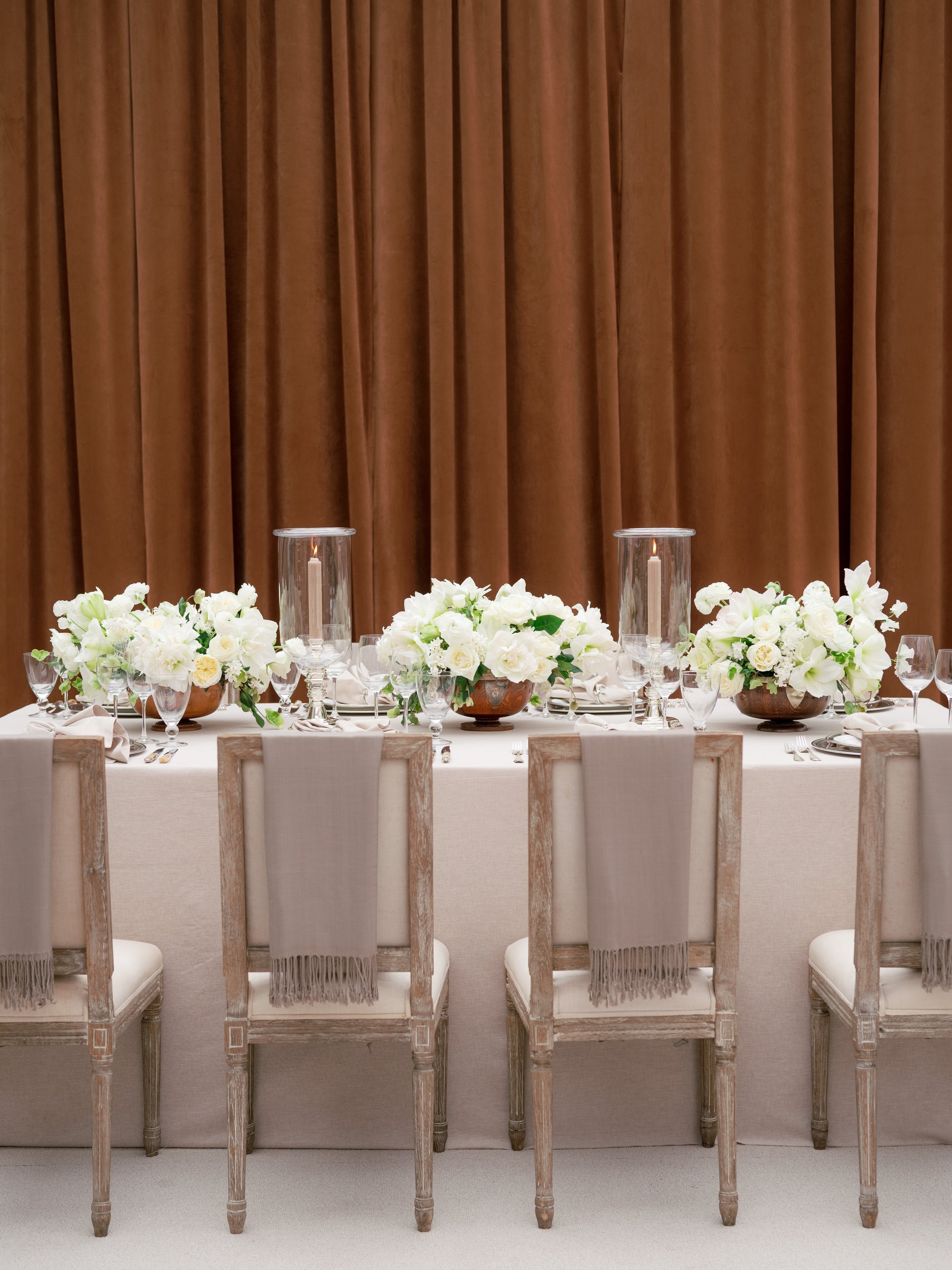 Elegant dining table decorated with white flowers, candles, and glassware, with a brown curtain in the background.