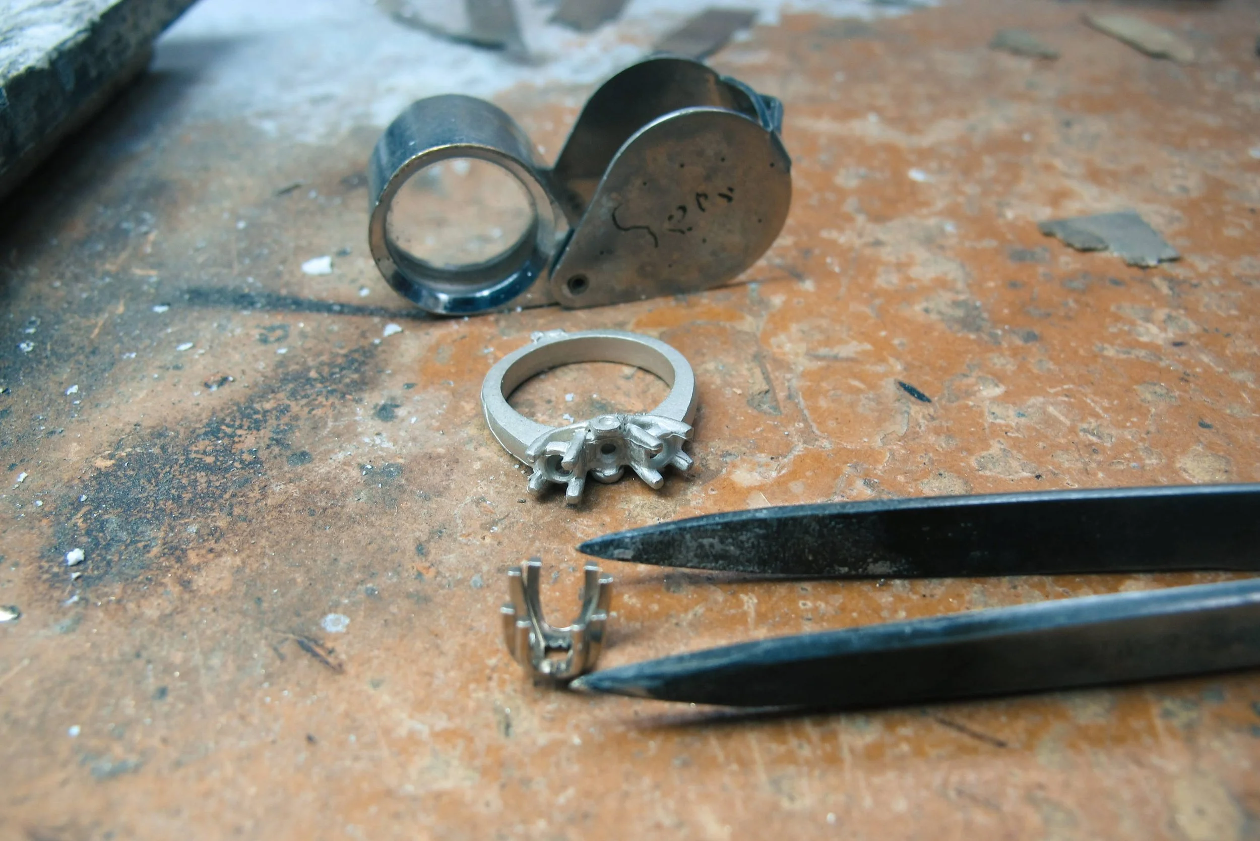 Unfinished ring mounting beside a loupe, tweezers and a loose setting on a workbench.