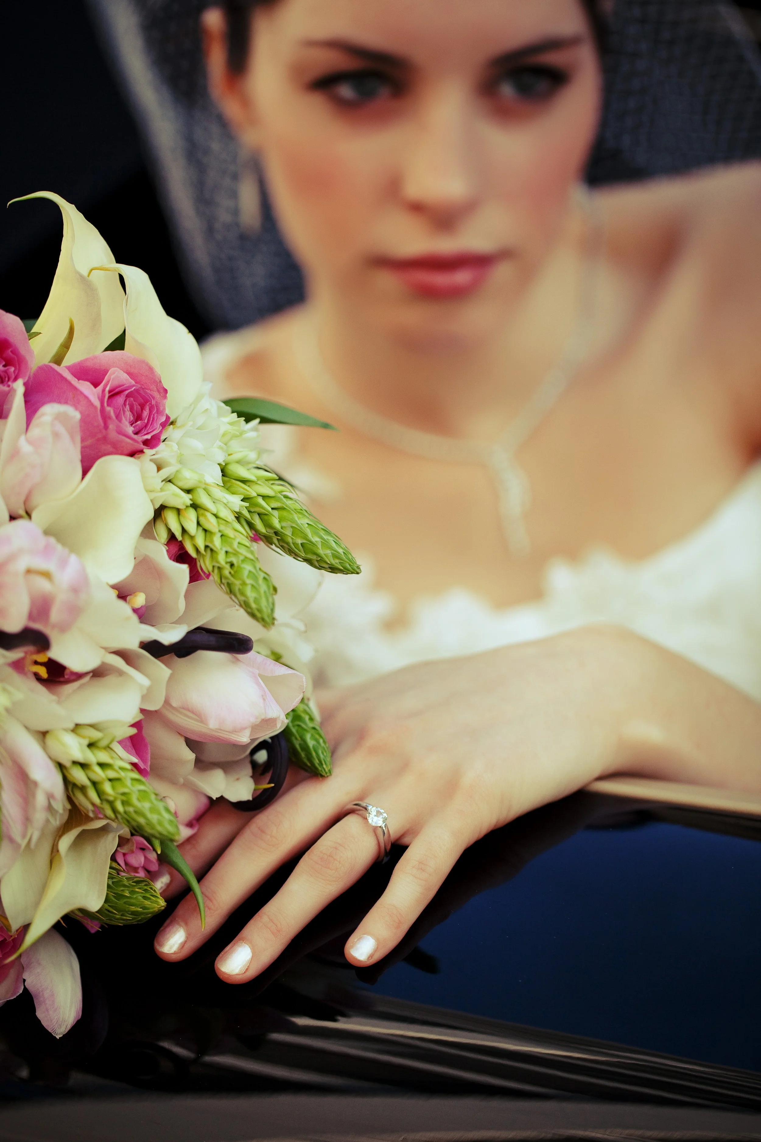 Bride wearing a clear solitaire ring while holding a pink and white bouquet beside a car.