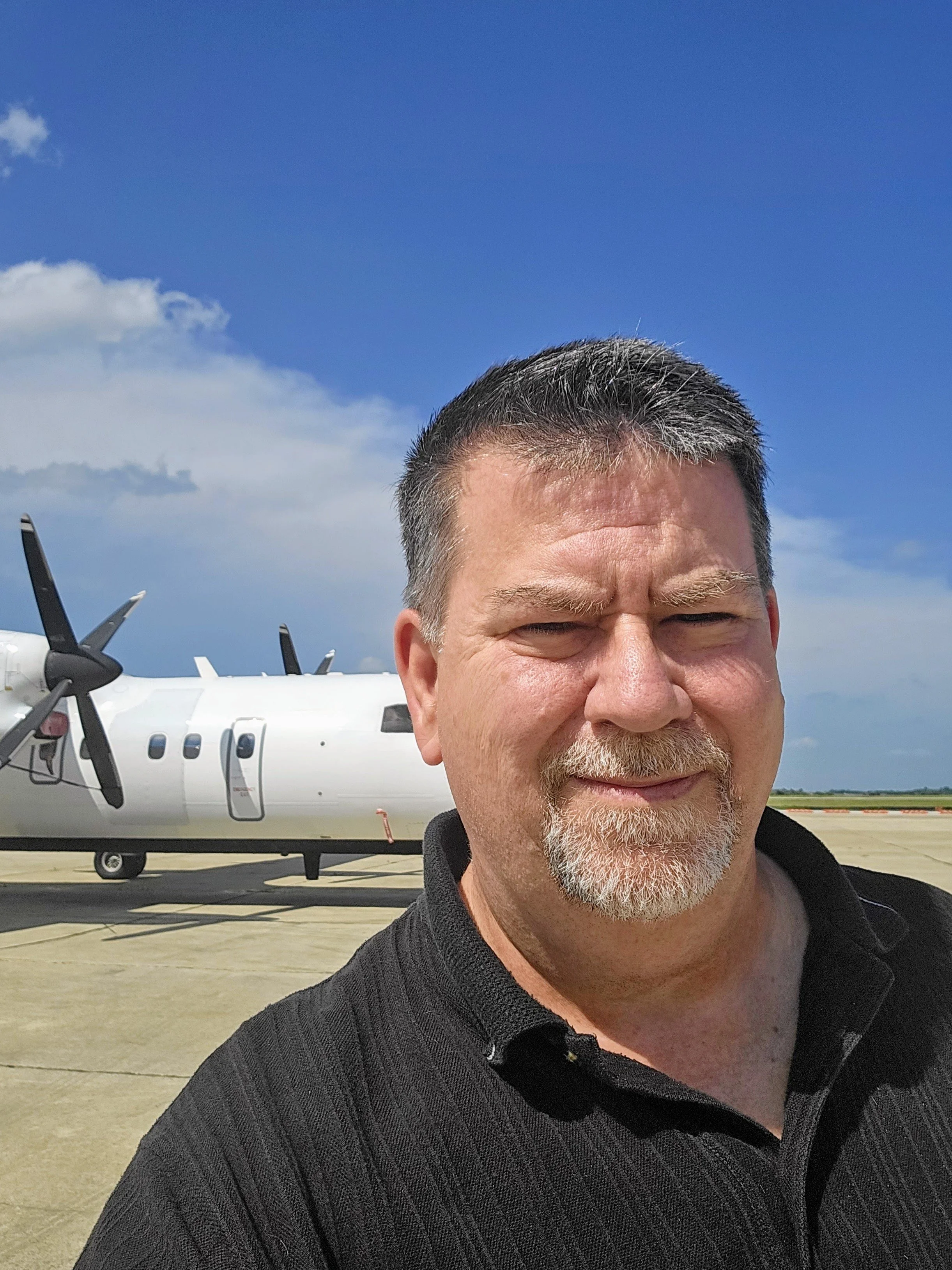 Close-up of a middle-aged man with gray hair and beard, smiling, standing outdoors on a sunny day with a blue sky and white clouds, in front of a white aircraft with propellers.