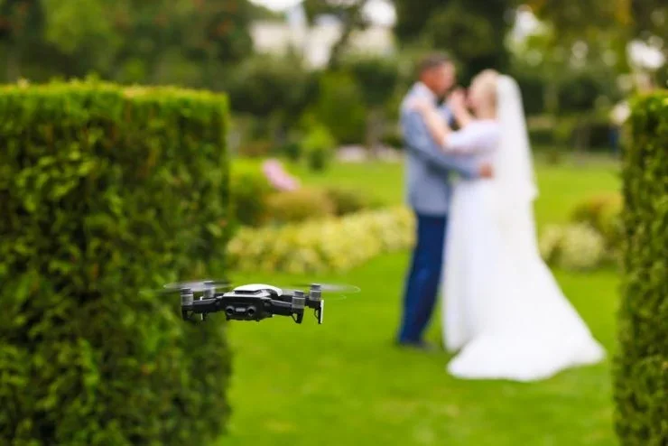 A drone flying in a park, with a couple dressed in wedding attire in the background.