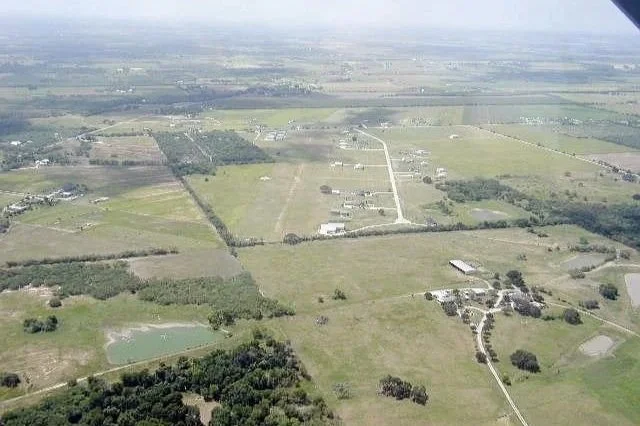 Aerial view of a rural landscape with open fields, scattered trees, dirt roads, and a few buildings under a cloudy sky.