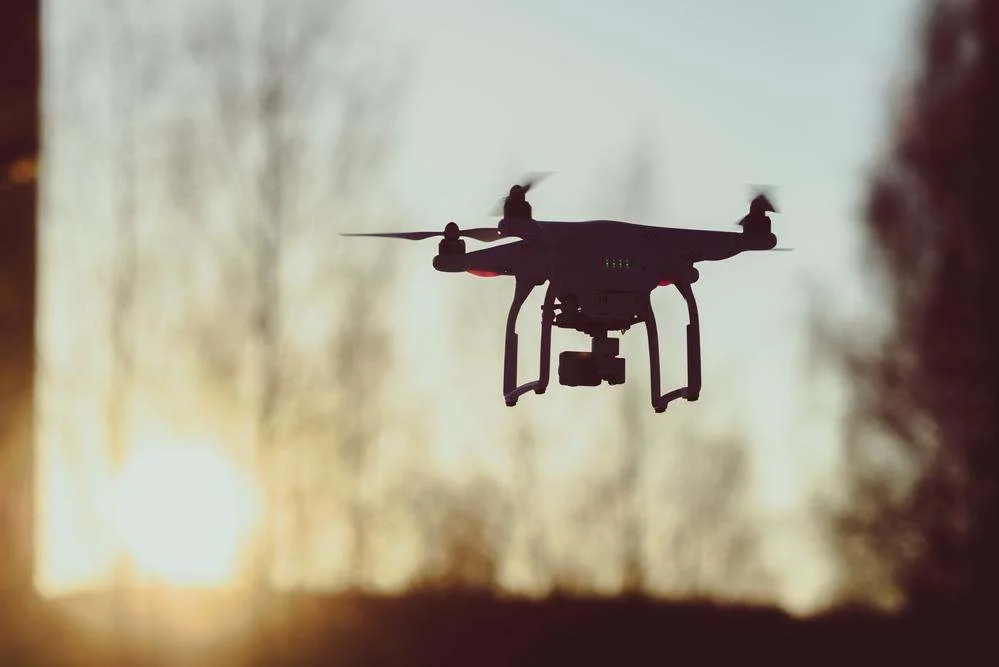 Silhouette of a drone flying outdoors during sunset or sunrise with a blurred background of trees.
