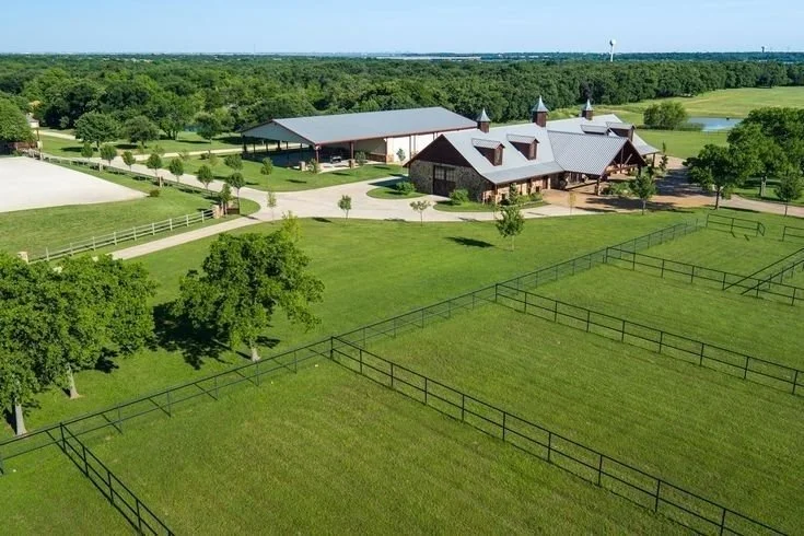 A large estate with a main house, a barn, and multiple fenced horse paddocks, surrounded by trees and open fields, under a clear blue sky.
