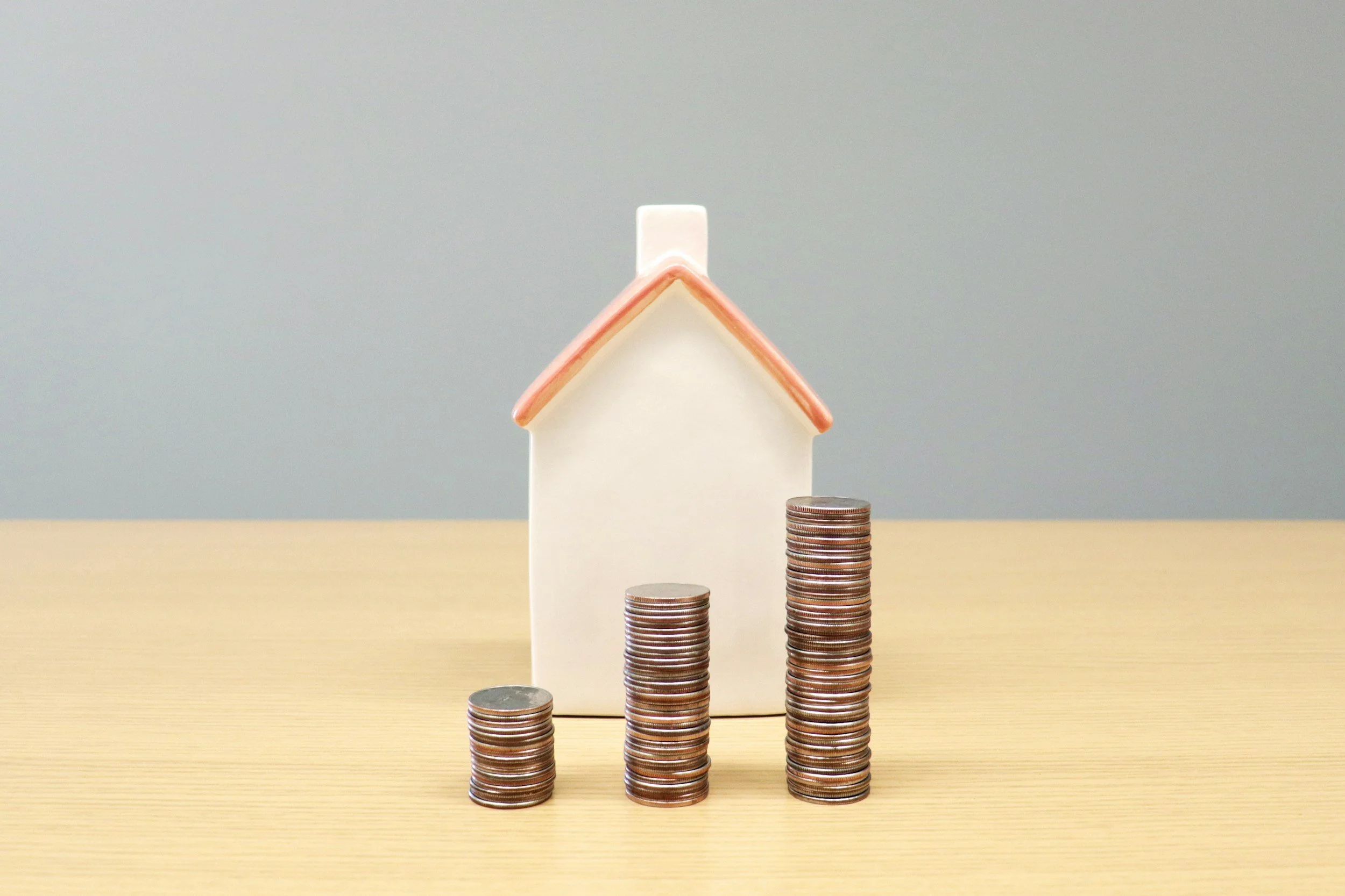 Stacks of coins increasing in height from left to right, with a small house model on top of the tallest stack, and a small white rectangular block at the house's peak.