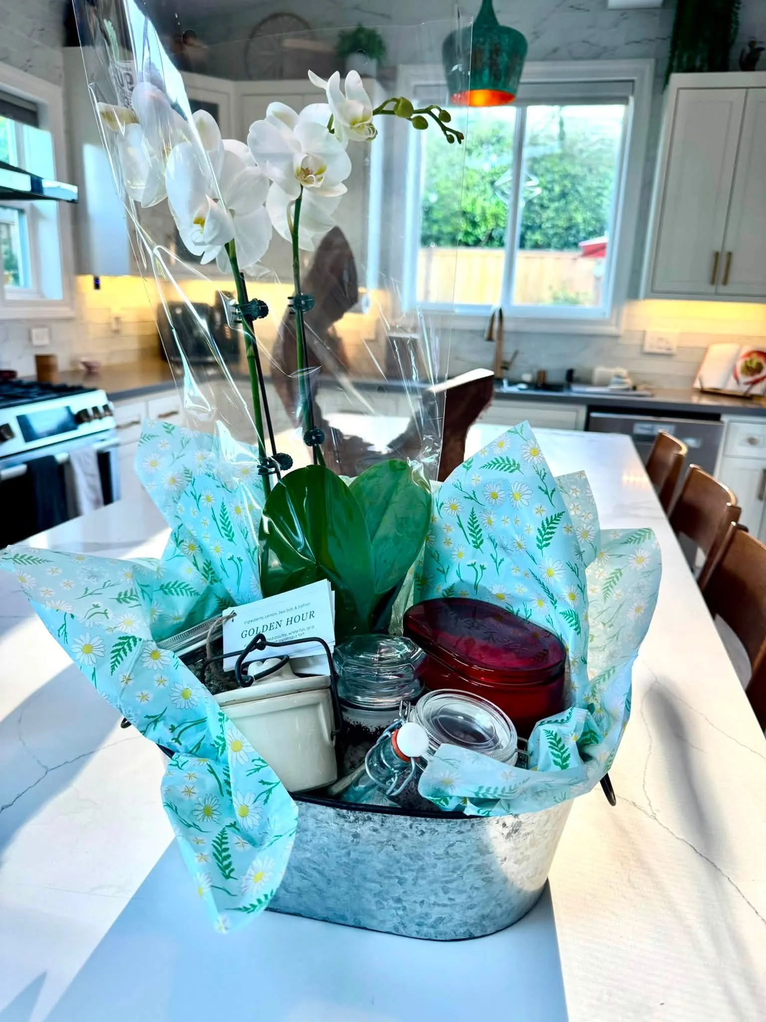 A white orchid in wrapping paper on a kitchen island next to a tray with small jars and a water bottle, with a modern kitchen background.