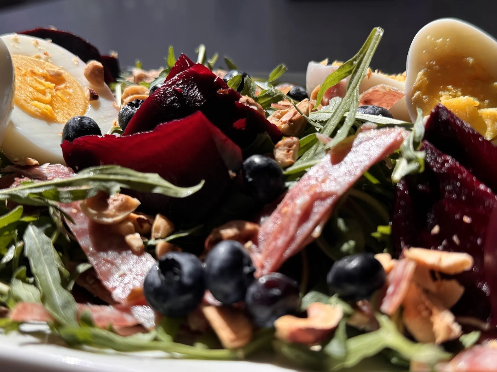Close-up of a salad with boiled eggs, blueberries, arugula, beet slices, chopped nuts, and thin slices of cured meat on a white plate.