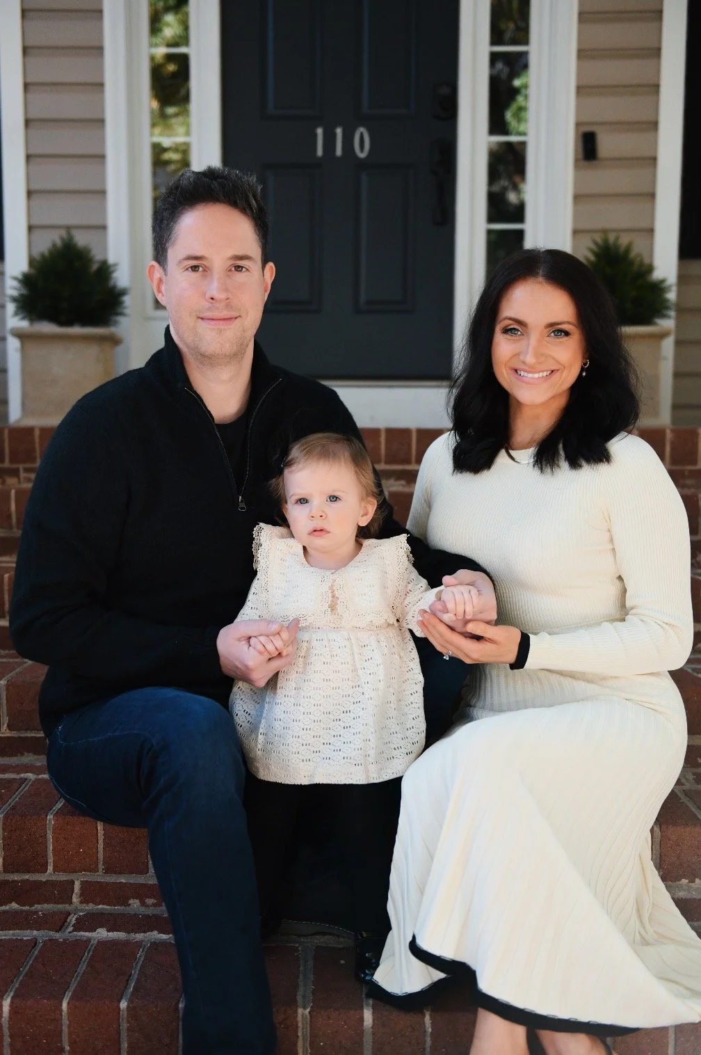 Family of three sitting on front steps of house, smiling. Man in black jacket, woman in cream dress, young girl in cream top.