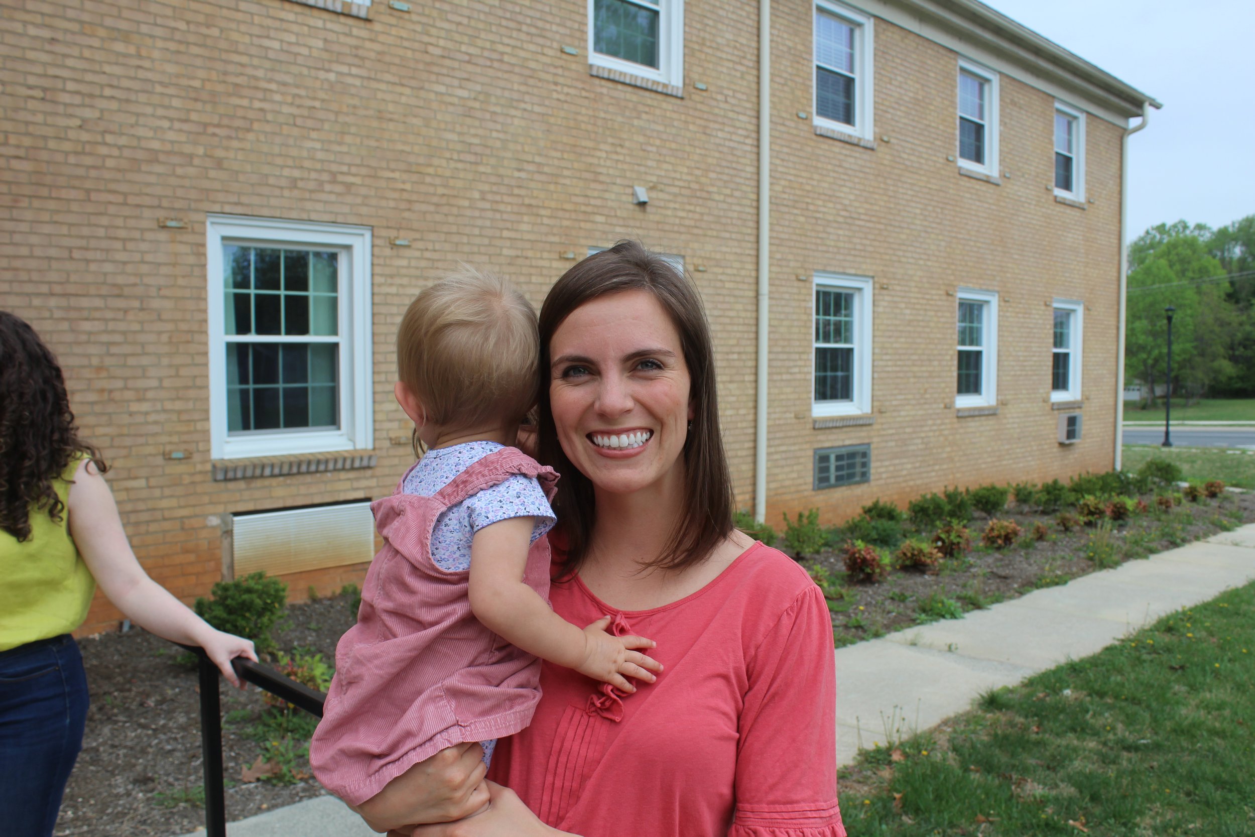 A woman holding a young girl in her arms outdoors in front of a brick building with multiple windows. The woman is smiling, and the girl is resting her head against the woman's face, partially obscured.