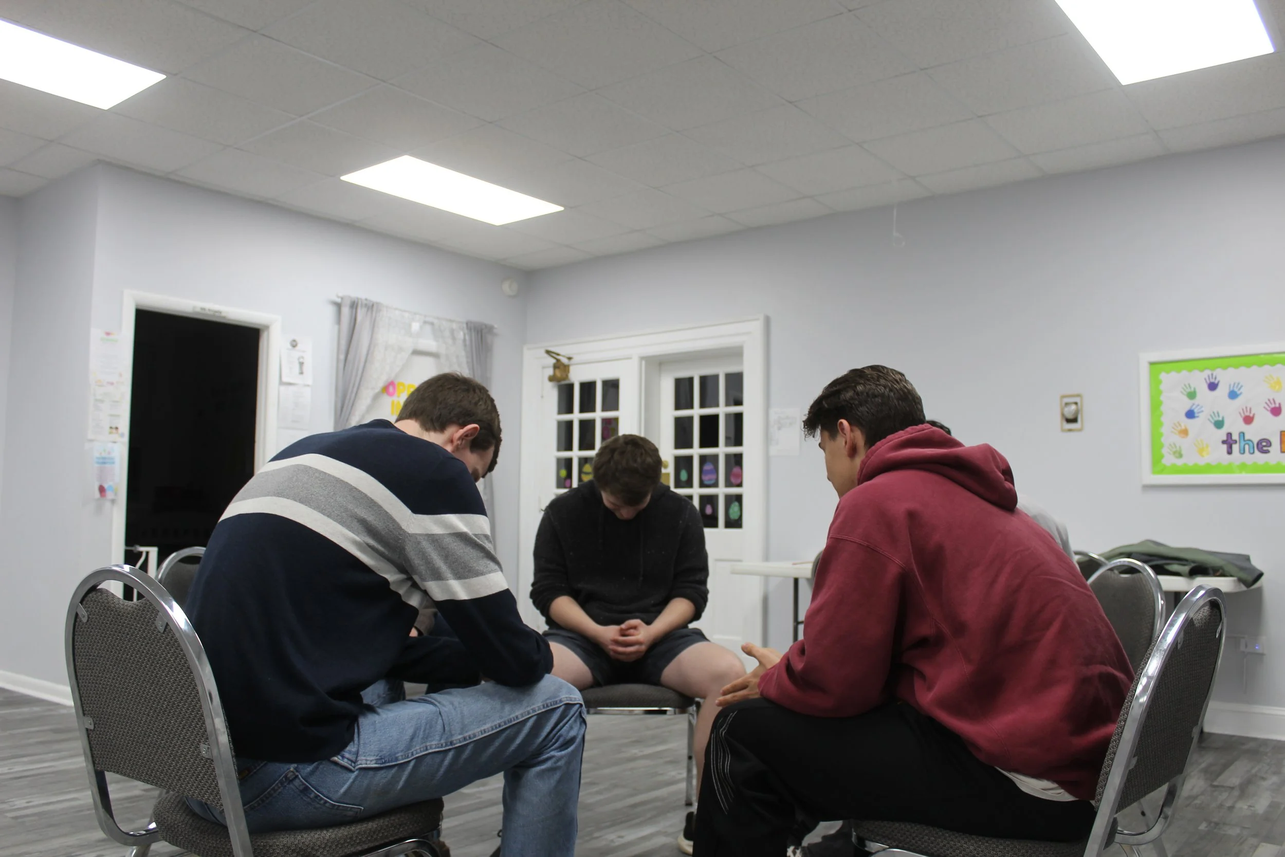 Four young men sitting on chairs with their heads bowed and hands clasped, praying in a room with gray walls and fluorescent ceiling lights.