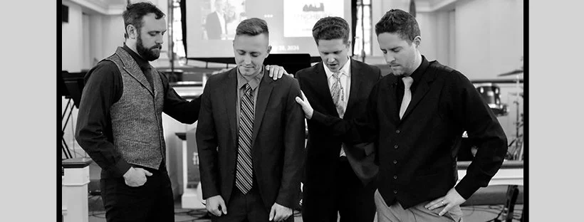 Four men in business attire standing together with heads bowed and hands on their shoulders in a professional setting.