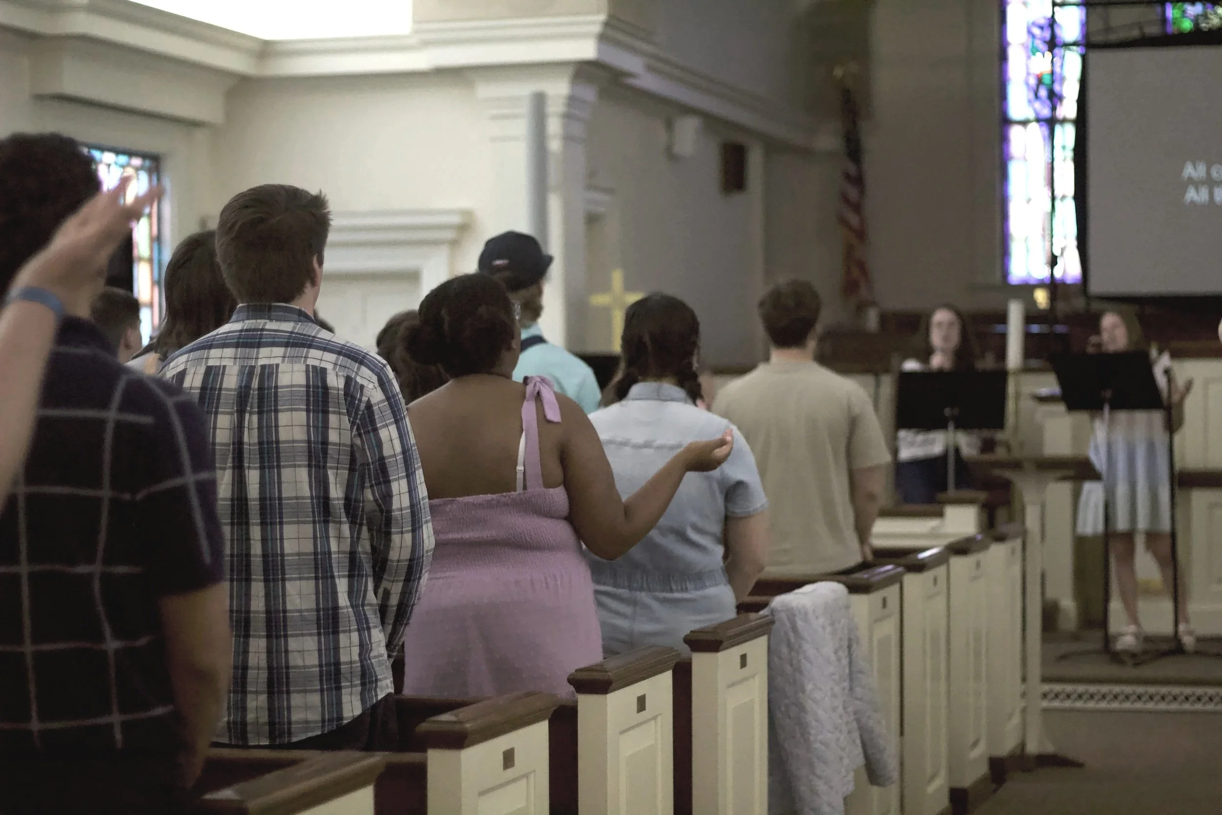 People standing inside a church, facing the altar, with some raising their hands in prayer or worship. There are stained glass windows and a large screen displaying song lyrics in the background.