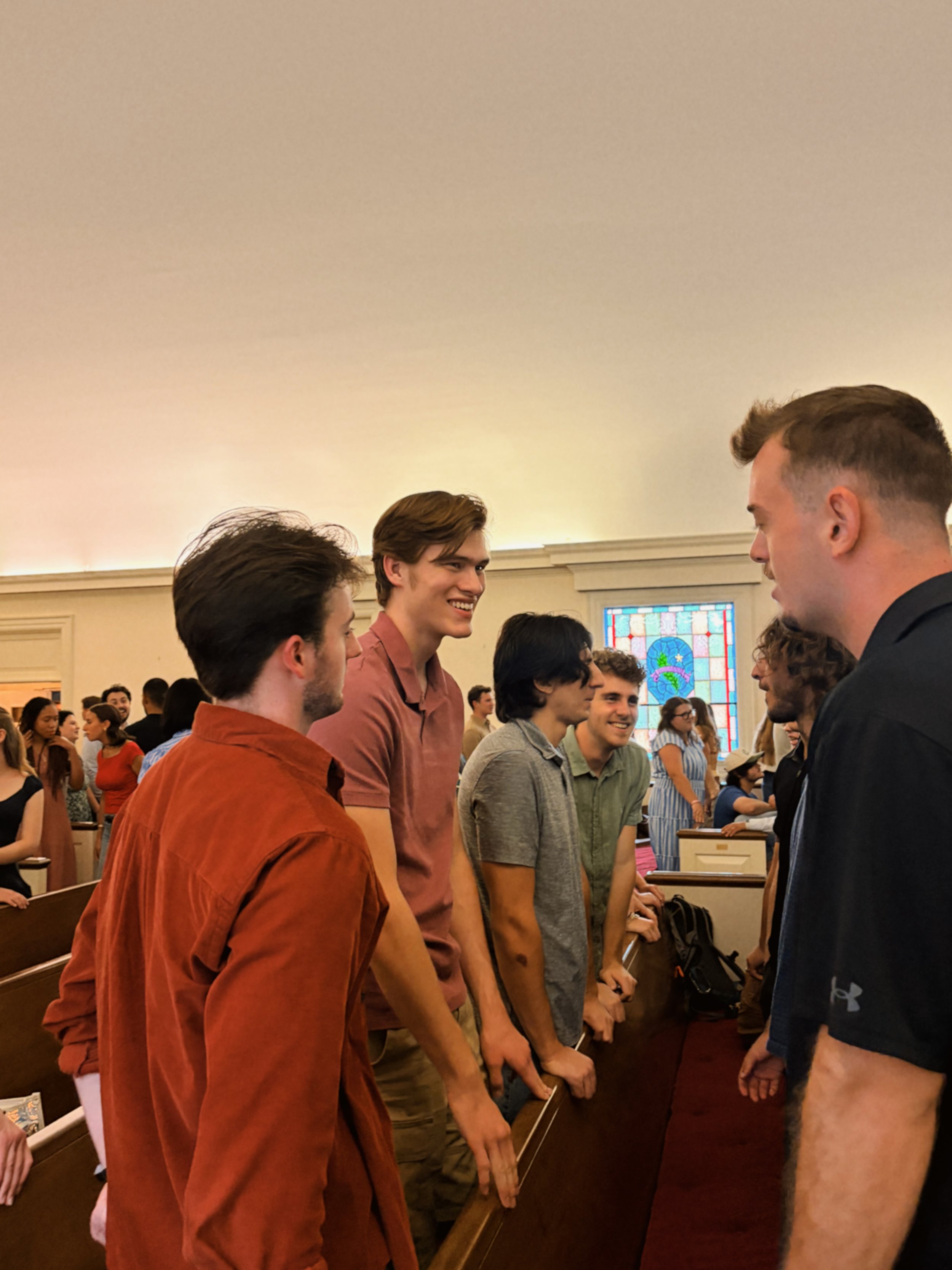 Young men and women standing face-to-face, engaged in conversation or greeting inside a church with stained glass windows.