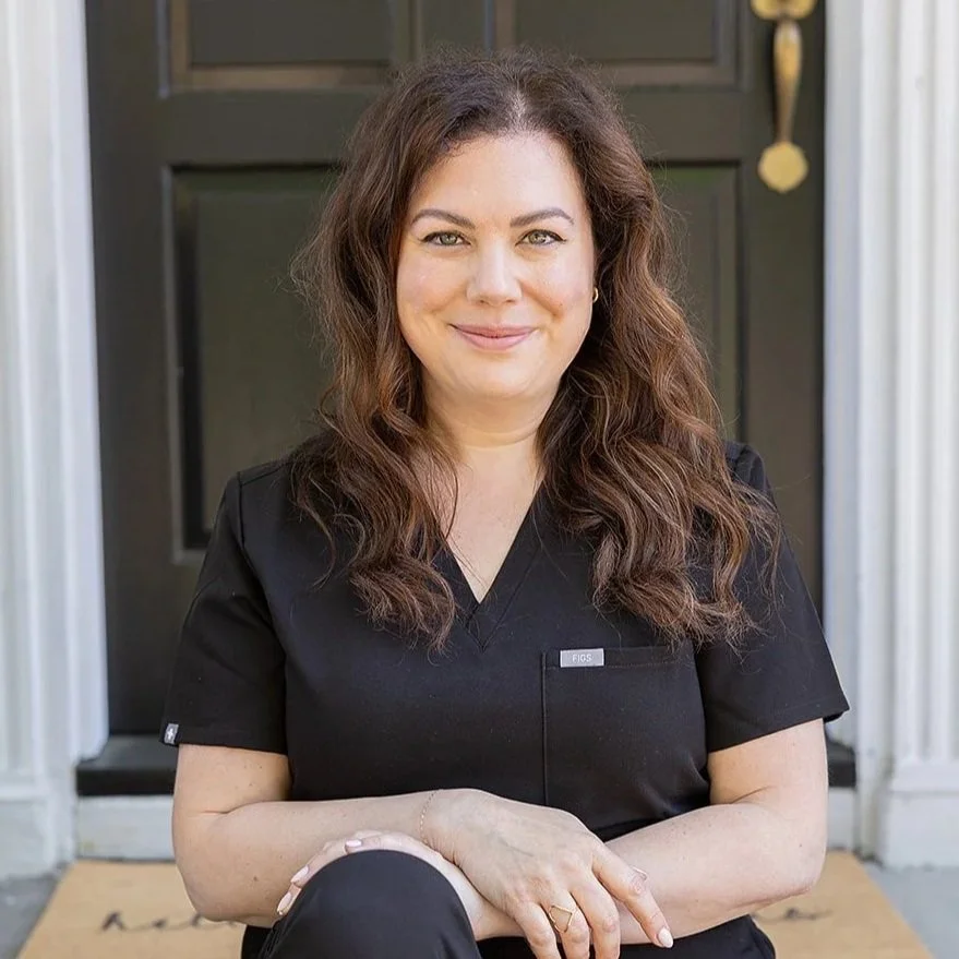 A woman with long, wavy brown hair, wearing black scrubs, sitting on a front porch in front of a black door. She is smiling and looking at the camera.