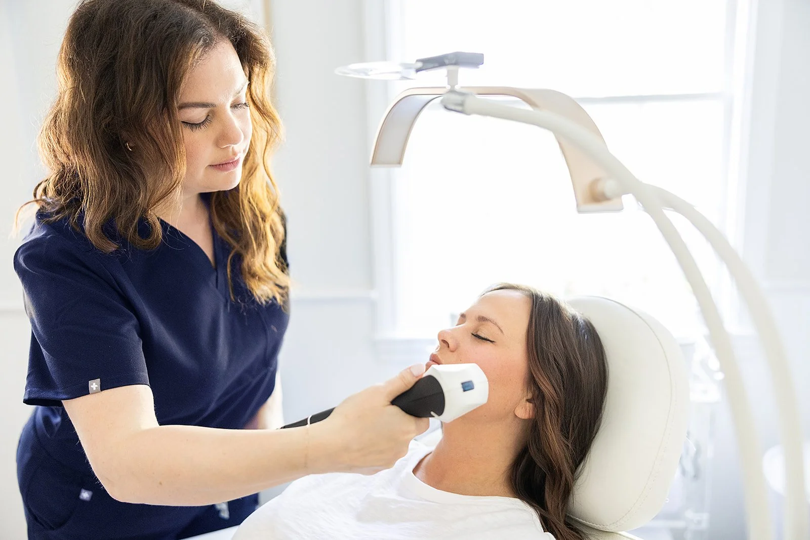 A woman receiving a facial treatment from a healthcare professional in a med spa with bright light and medical equipment.